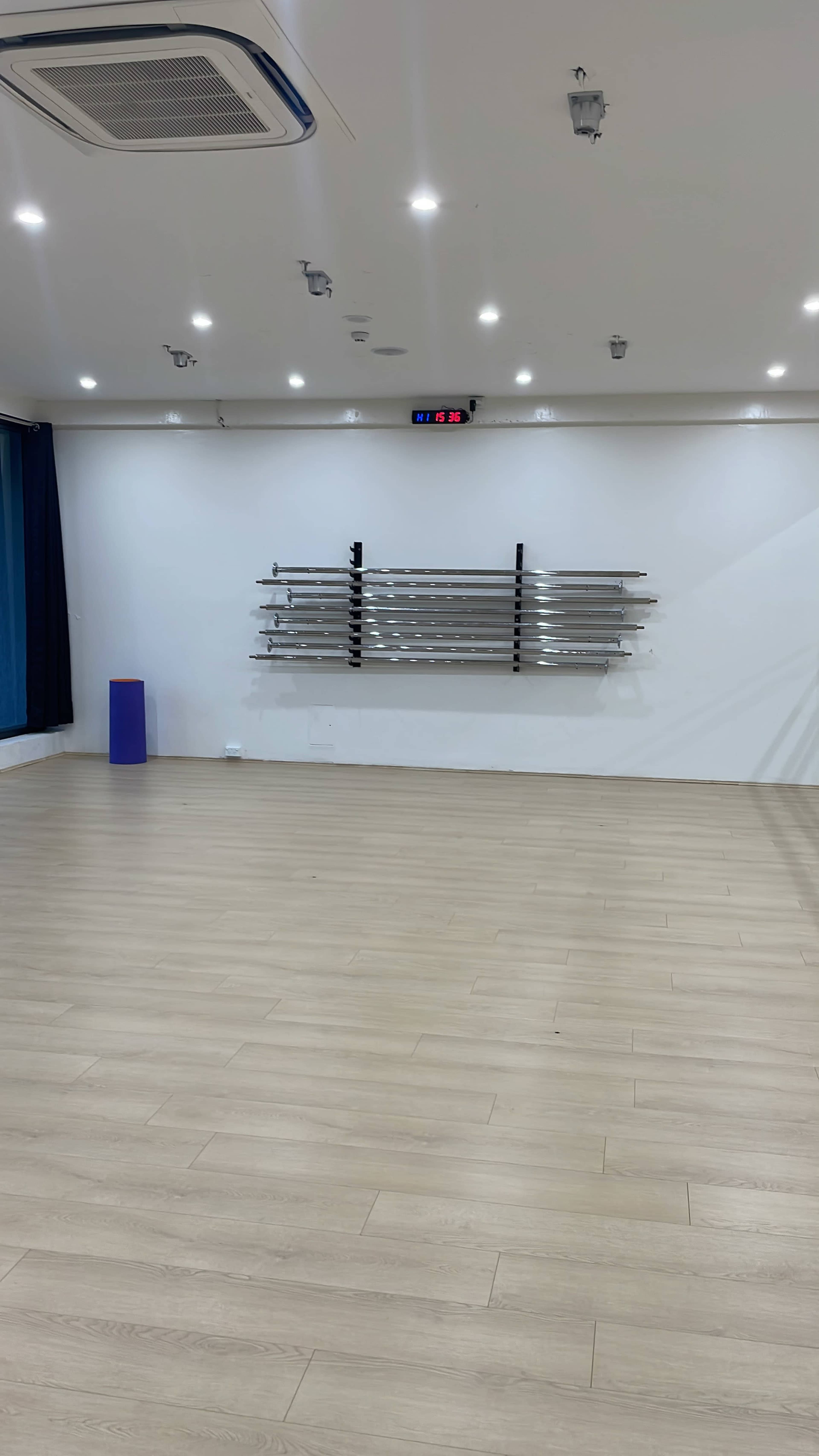A spacious, well-lit room with wooden flooring, white walls, a blue curtain, and a wall-mounted shelf holding several metal rods.