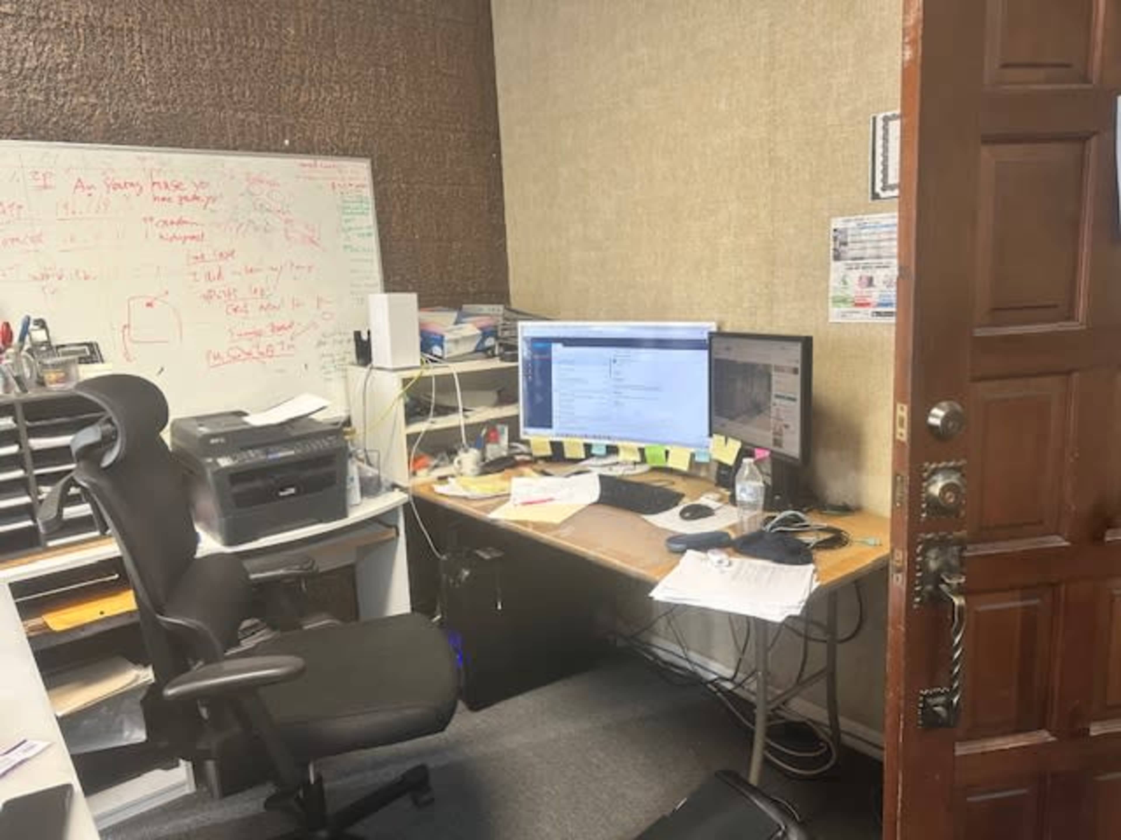 The image shows a small office with a desk, computer, printer, and a wall covered in whiteboard and cluttered notes.