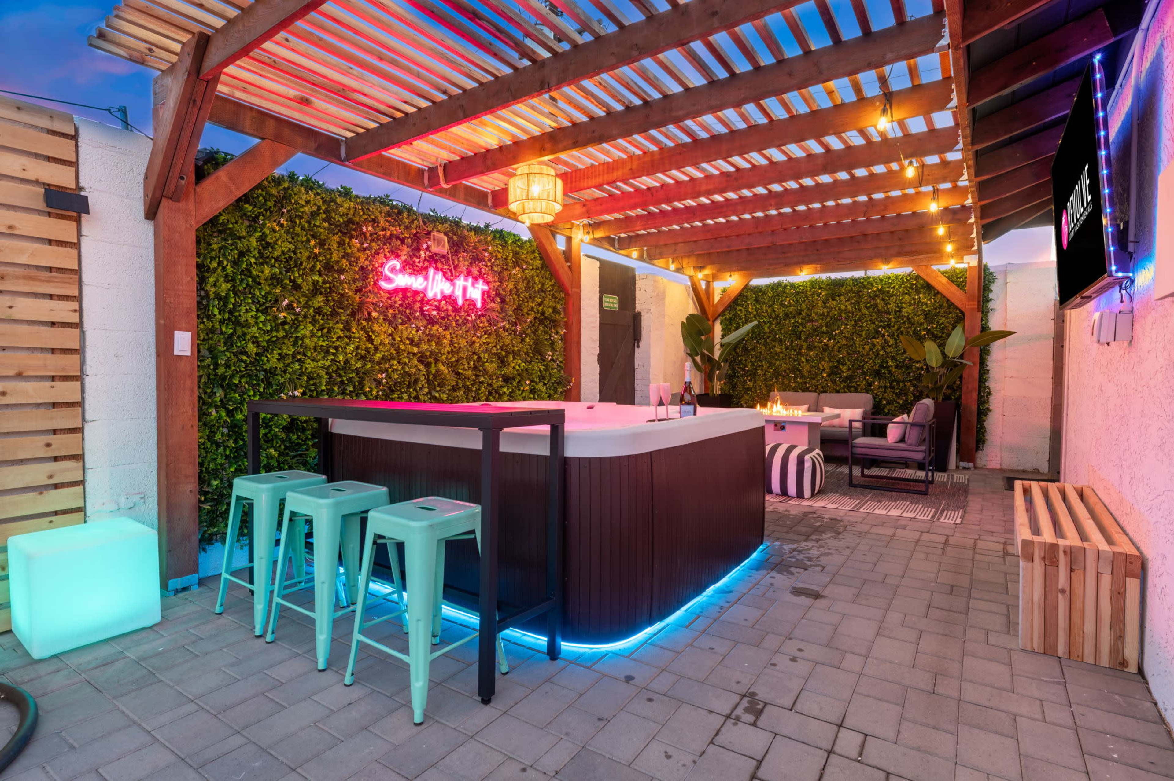 The image shows a backyard setting featuring a bar area with high stools, surrounded by greenery and illuminated by colorful lights.