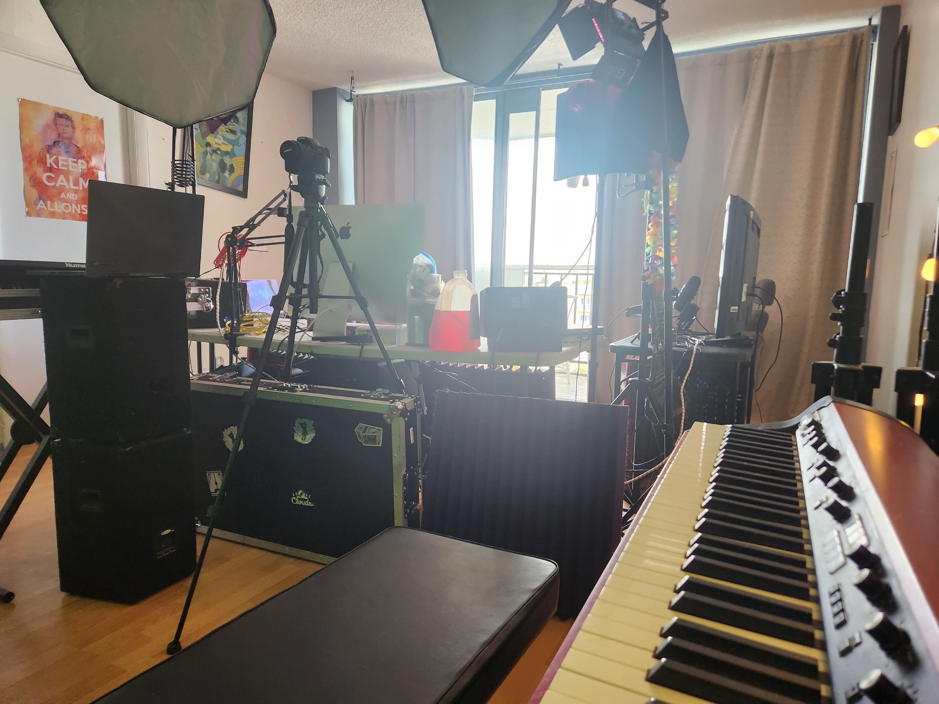 A workspace setup features a variety of equipment including cameras, computers, and a keyboard, with large windows in the background allowing natural light to enter.