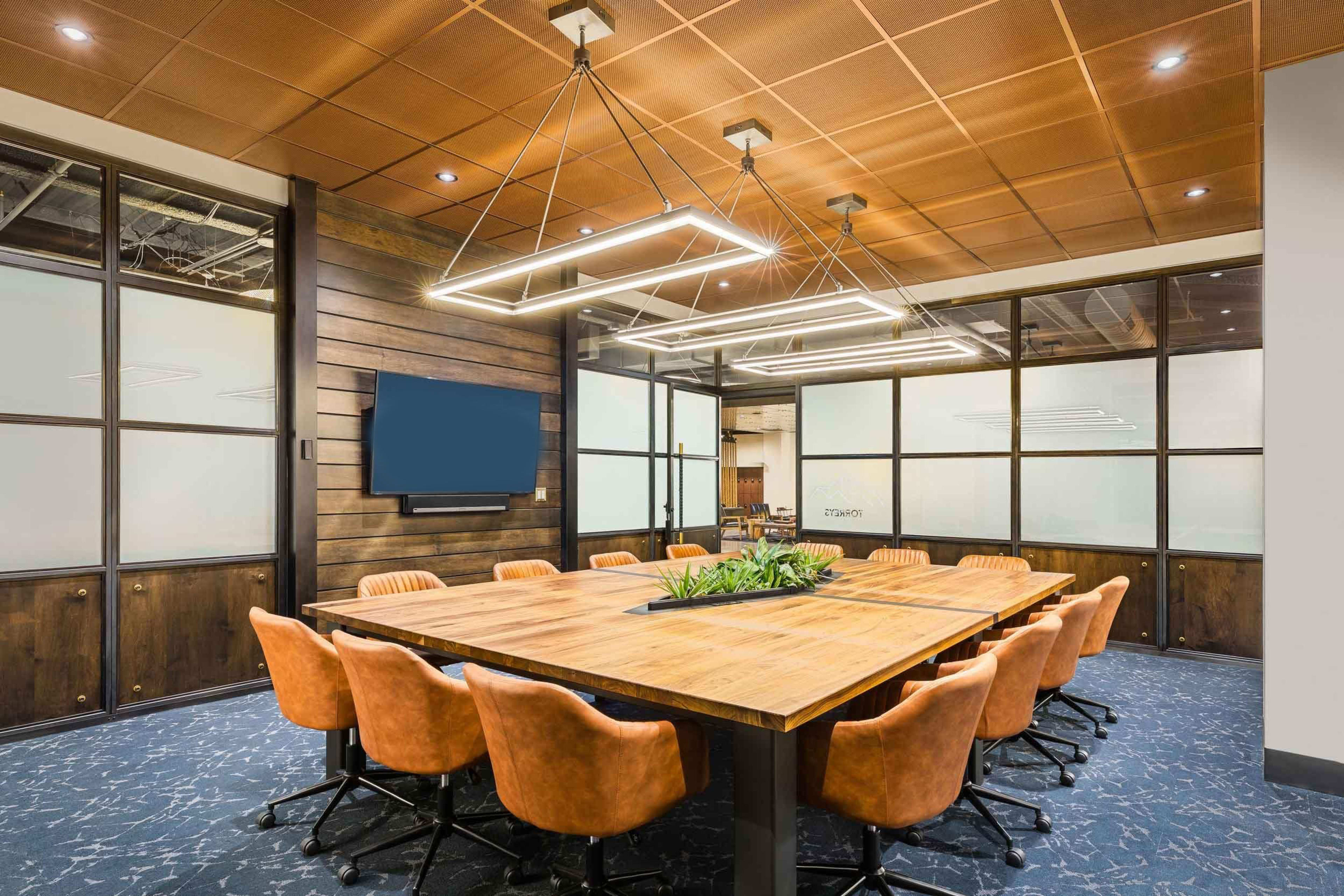 A modern conference room with a large wooden table surrounded by orange ergonomic chairs and a television mounted on the wall.