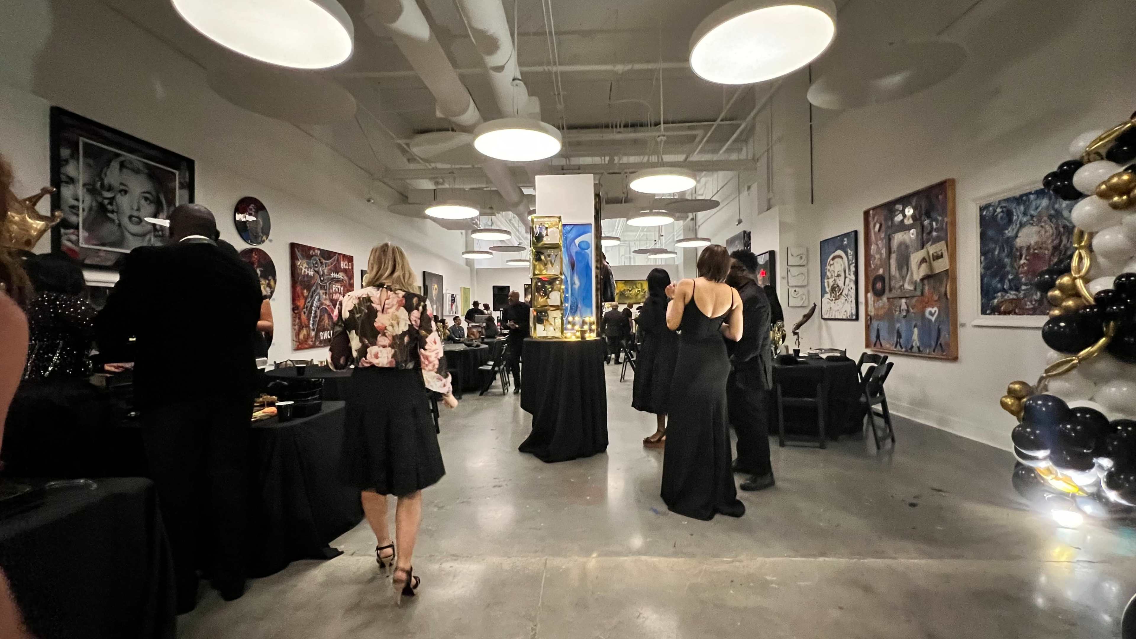 A group of elegantly dressed guests mingle in a gallery with artistic displays and black tables set for an event.