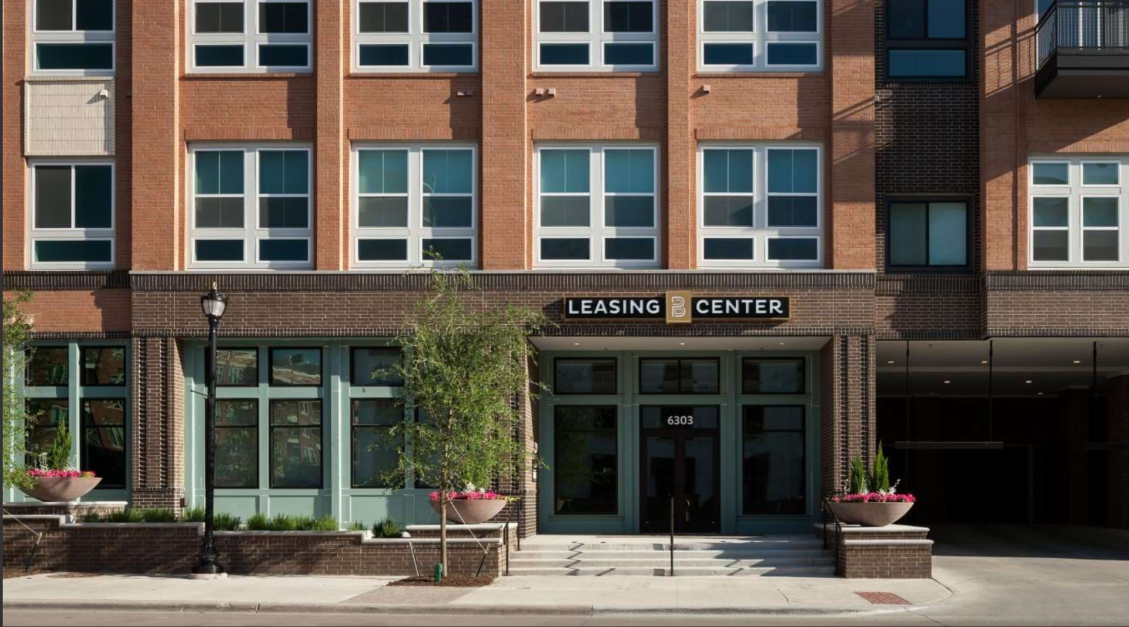 The image shows a modern multi-story building with a prominent leasing center entrance featuring large windows and planters on either side.