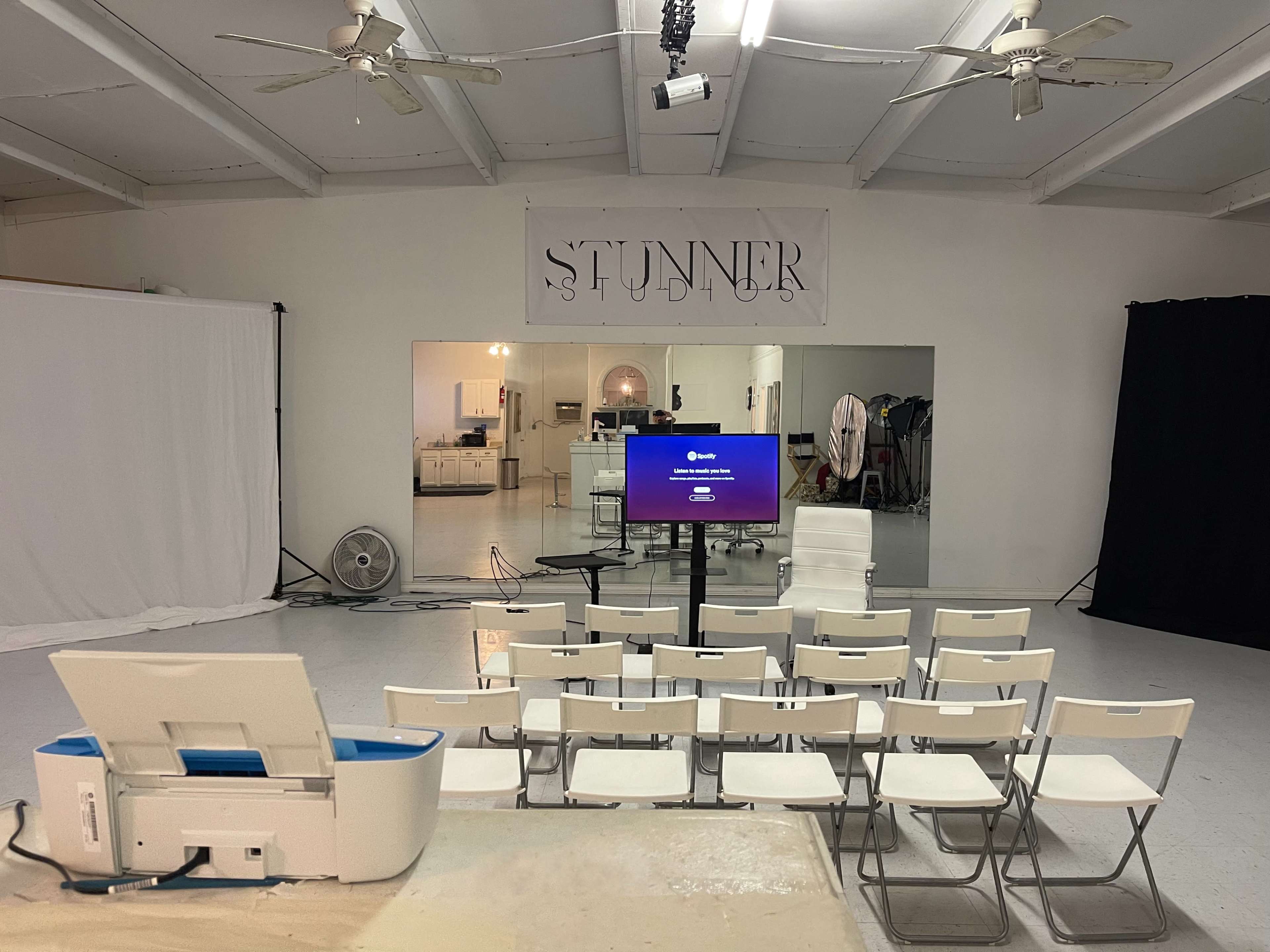 The image shows a minimalist studio setup with rows of chairs facing a screen, framed by white walls and a large banner stating "STUNNER."
