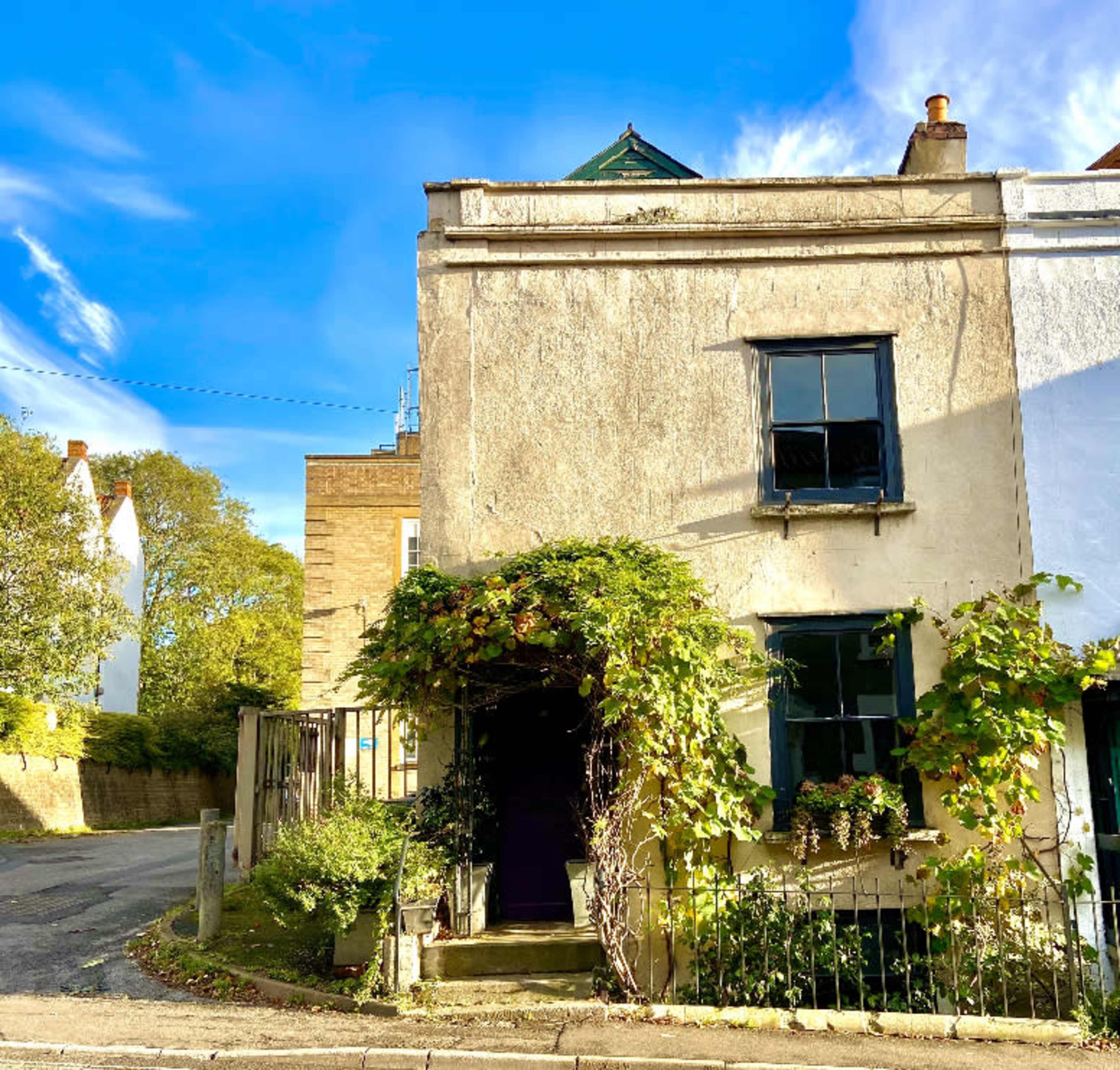 A weathered house with a green vine covering the entrance is located at the corner of a quiet street under a blue sky.