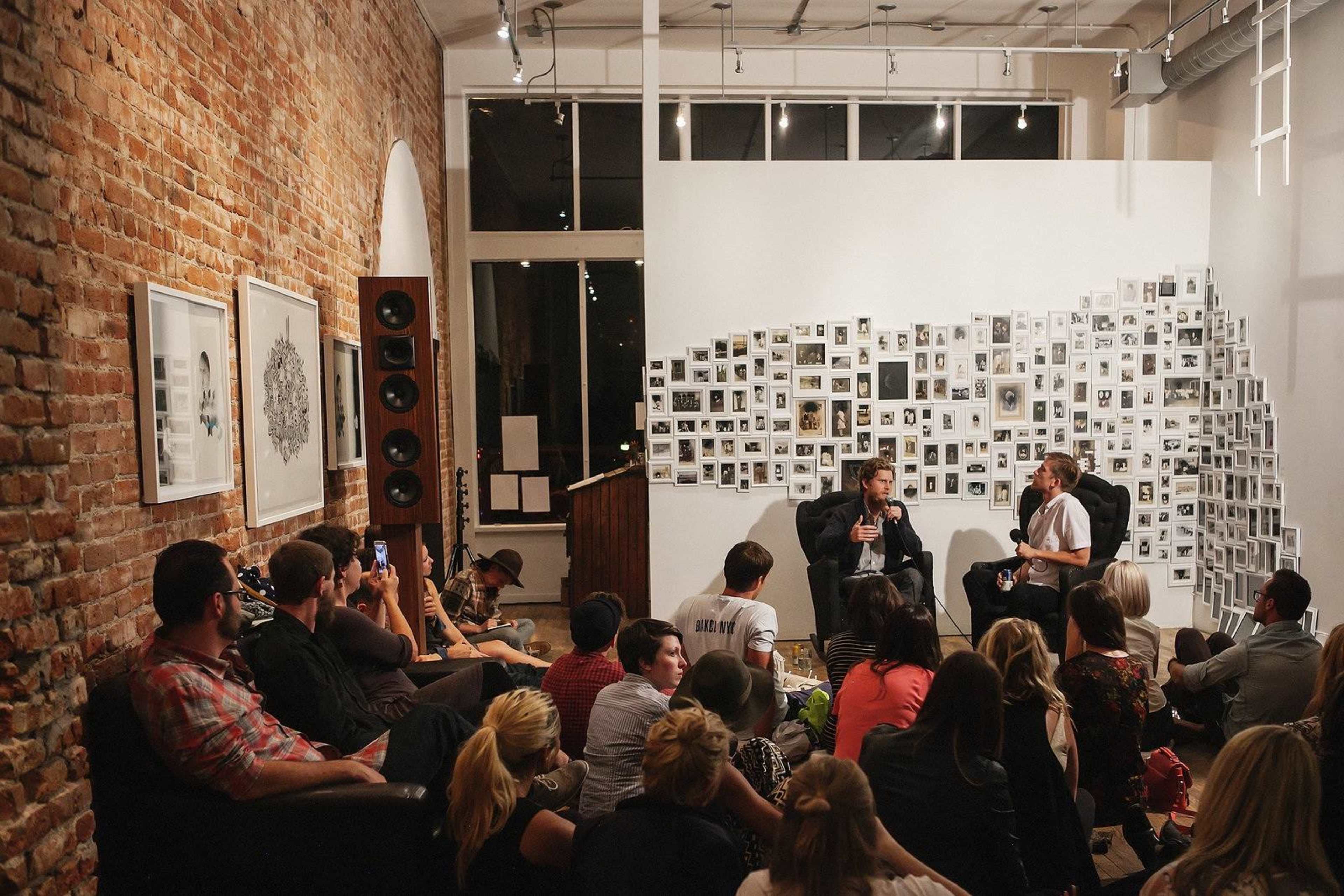 A small crowd sits on the floor and chairs in a gallery space, while two speakers engage in a discussion near a wall covered with numerous photographs.