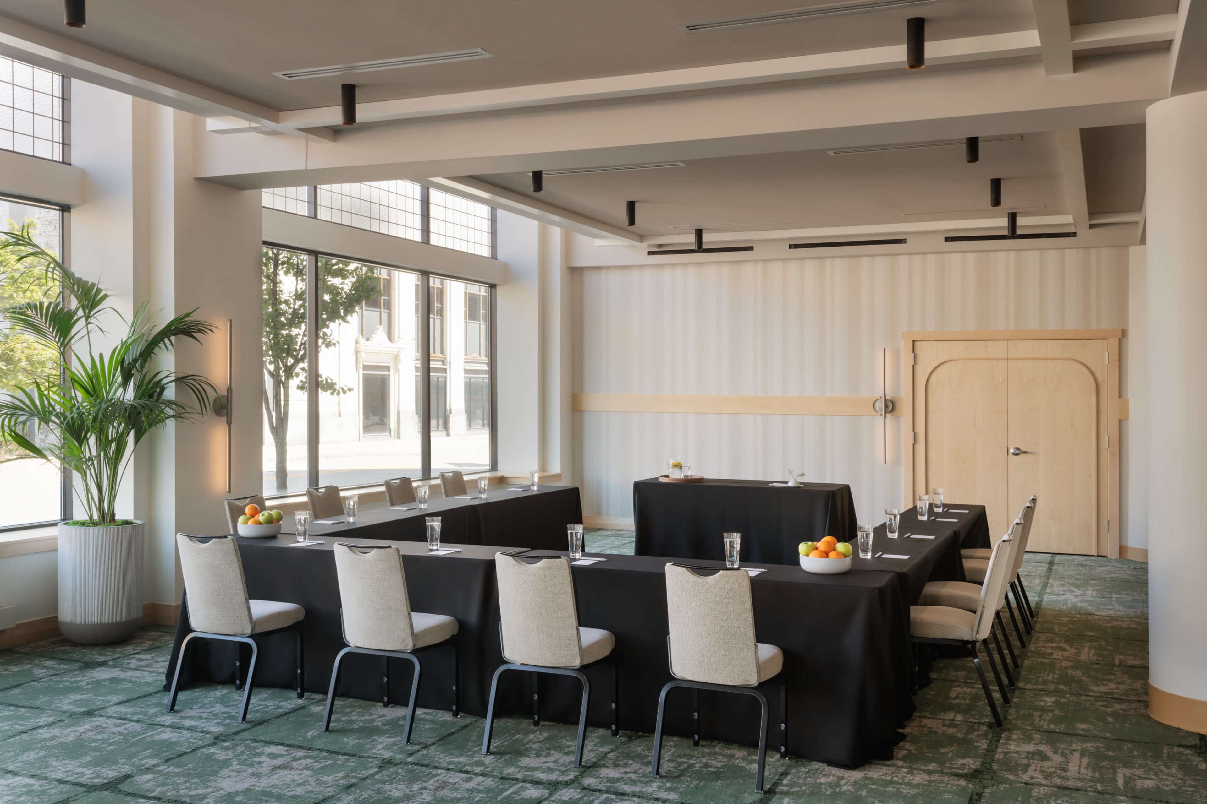 A modern conference room features a long table with fruit and water bottles, surrounded by chairs, and large windows offering natural light.