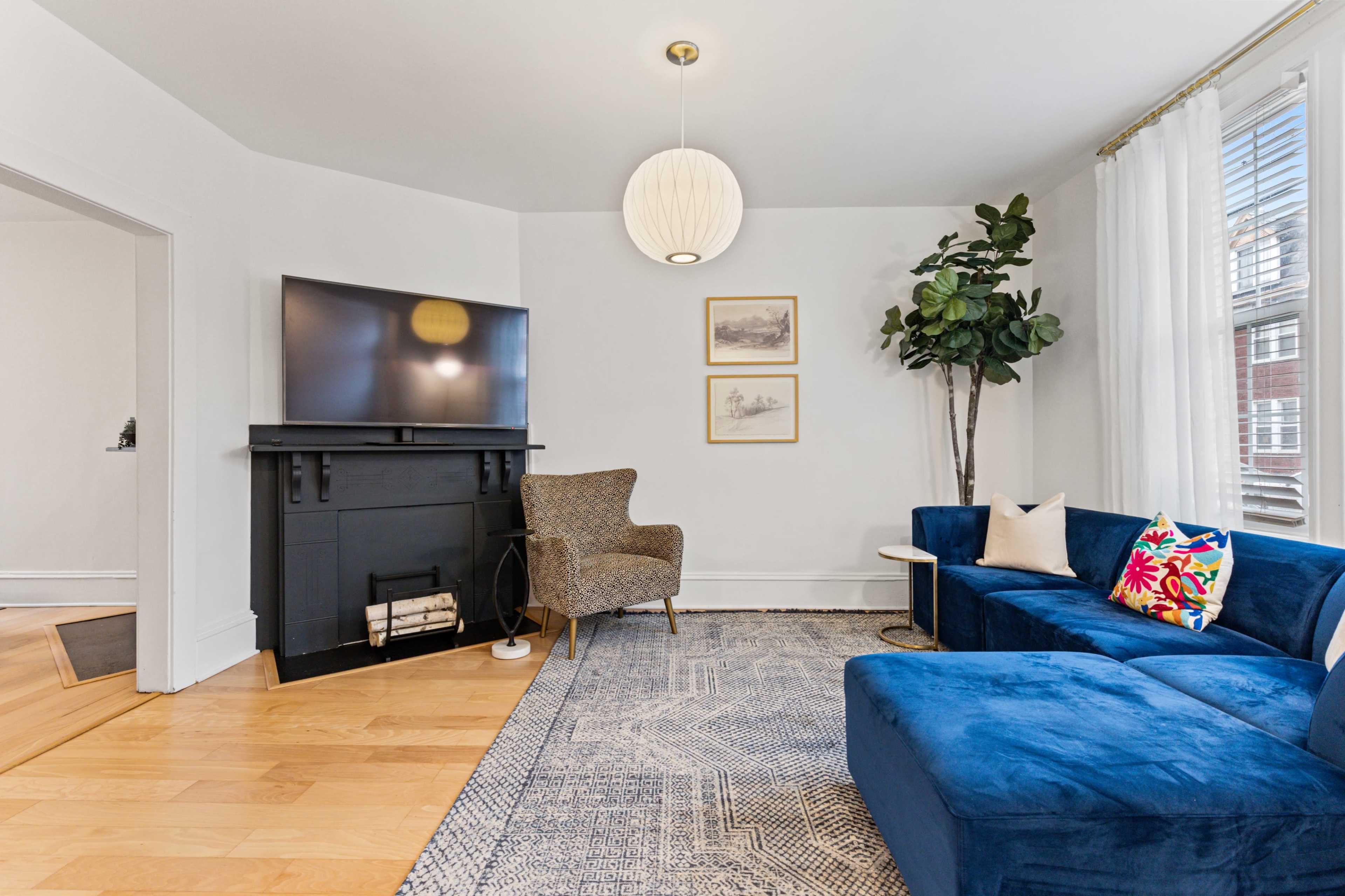 A living room featuring a blue sofa, an armchair, a fireplace with logs, and a large television mounted on the wall.
