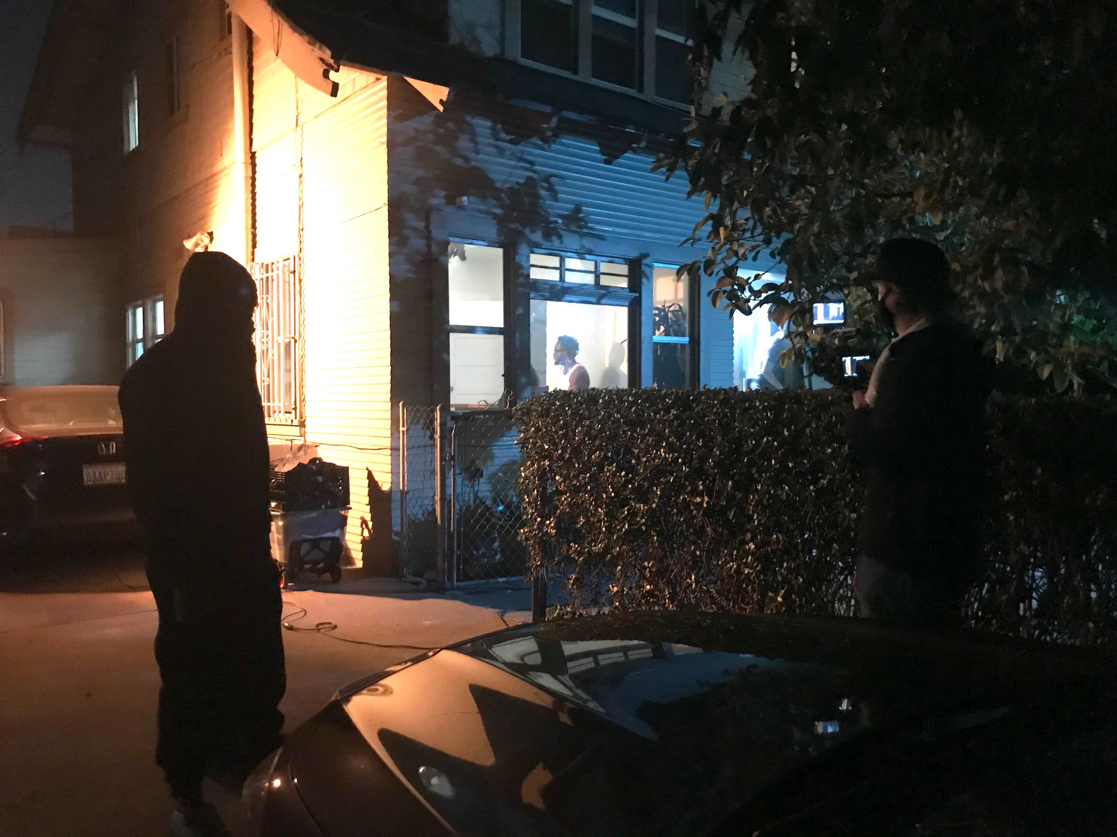 A person in a hooded sweatshirt stands outside a house while another person inside is filmed by a man holding a camera.