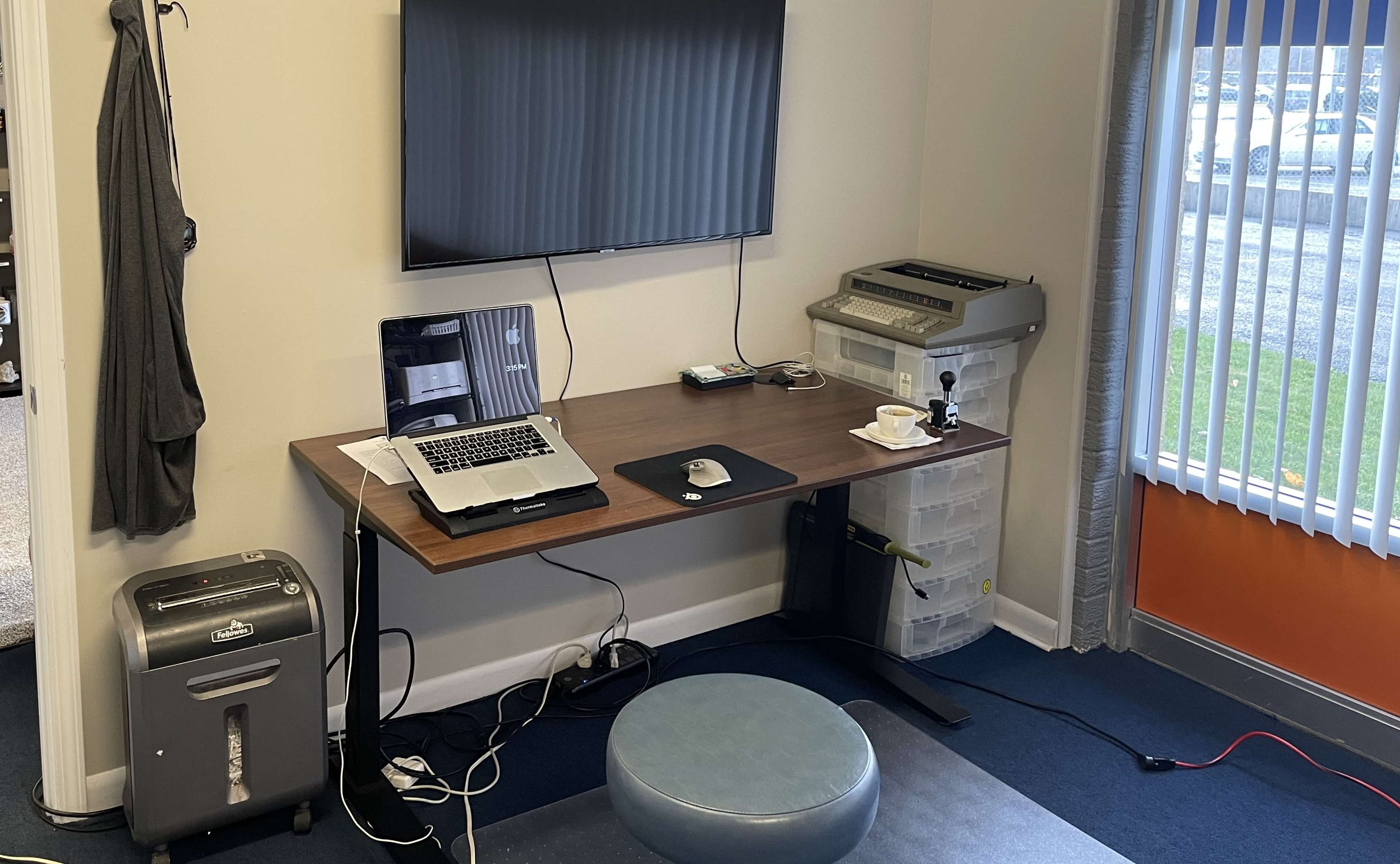 A modern workspace features a wooden desk with a laptop, printer, and storage container, accompanied by a small stool and a television mounted on the wall.