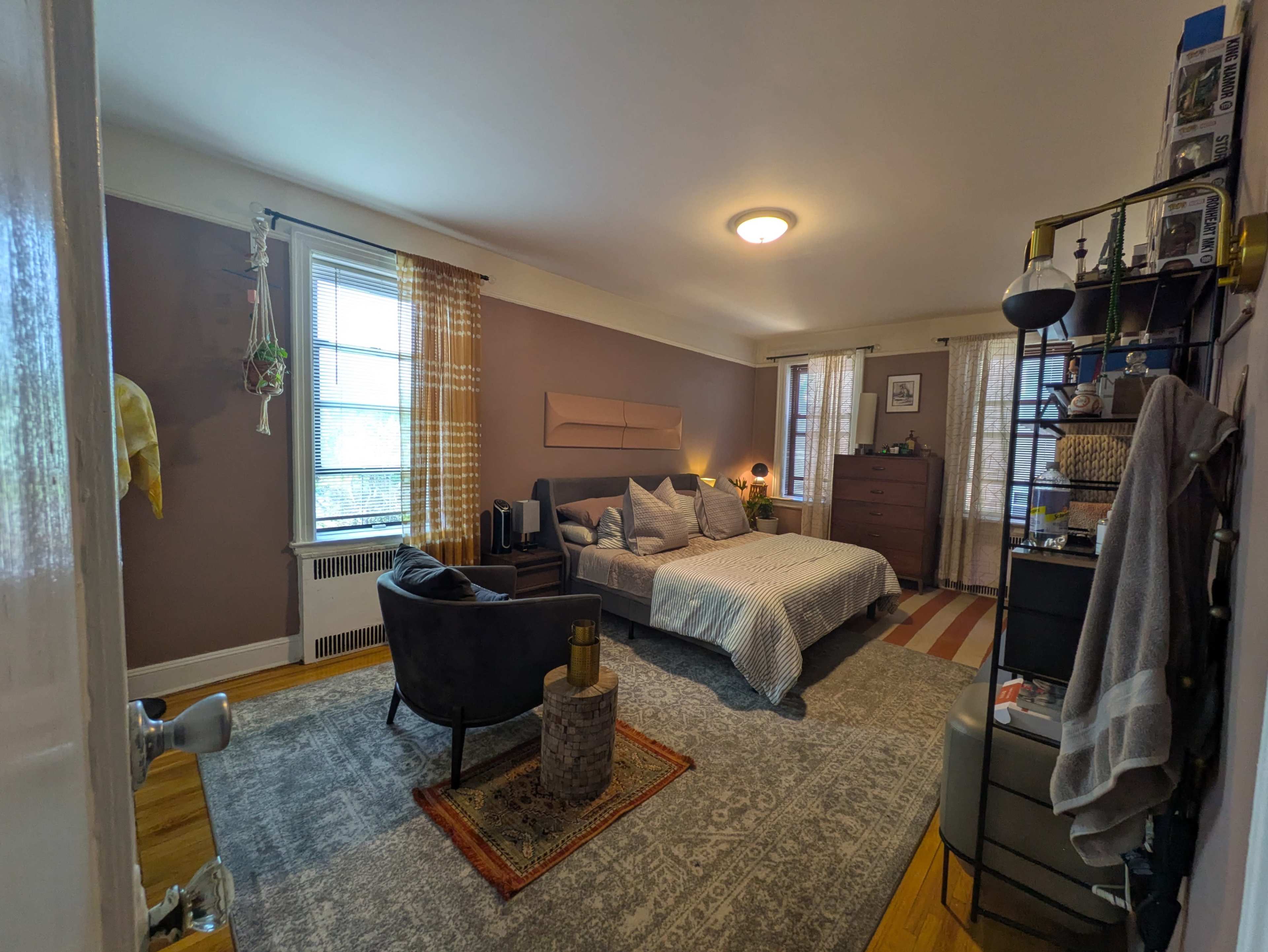 The image shows a well-furnished bedroom featuring a bed, dresser, and a sitting area with a rug and decorative items.
