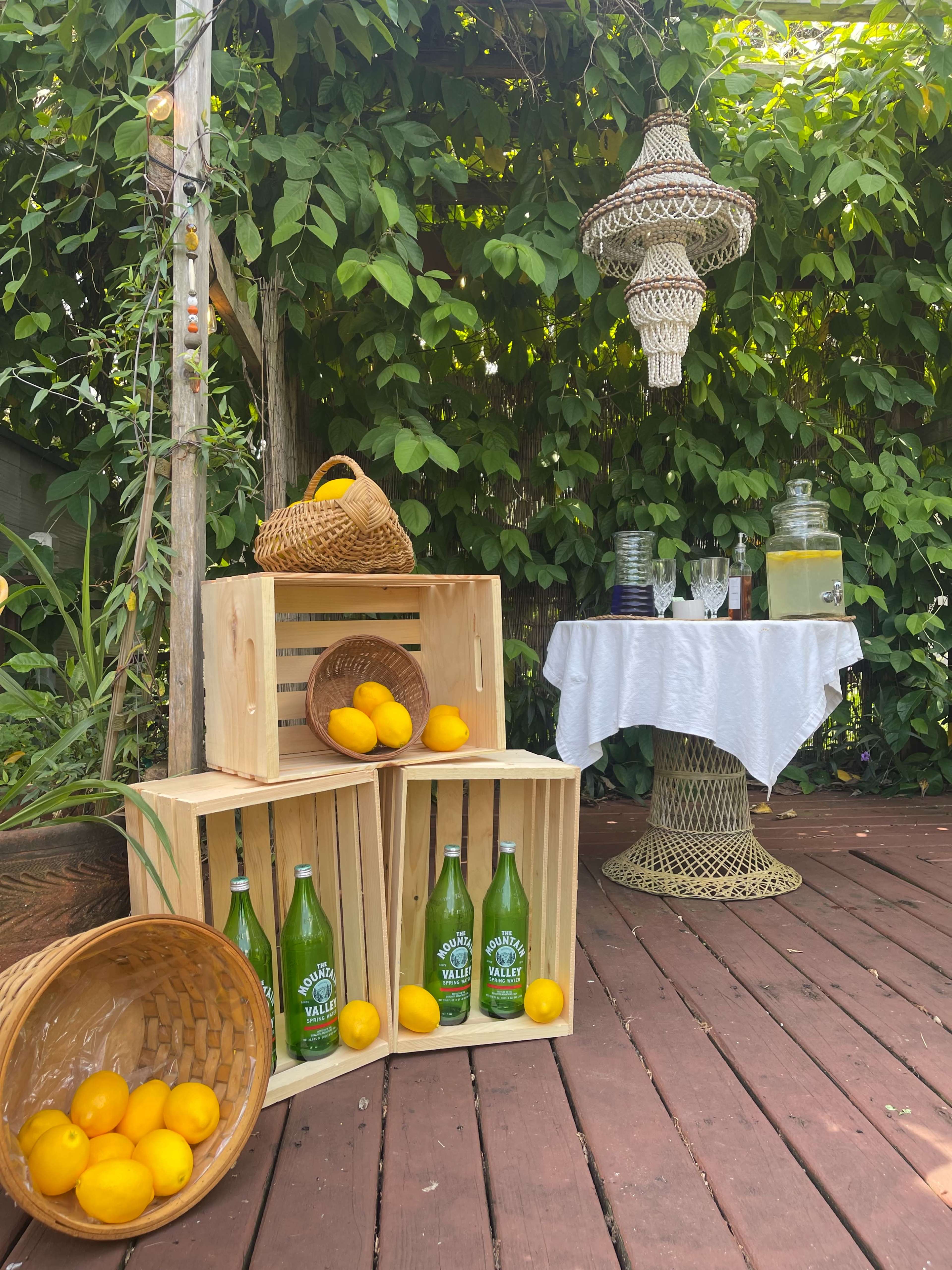 The scene features wooden crates filled with lemons beside green glass bottles and a table draped in white fabric, all set against a backdrop of lush greenery.