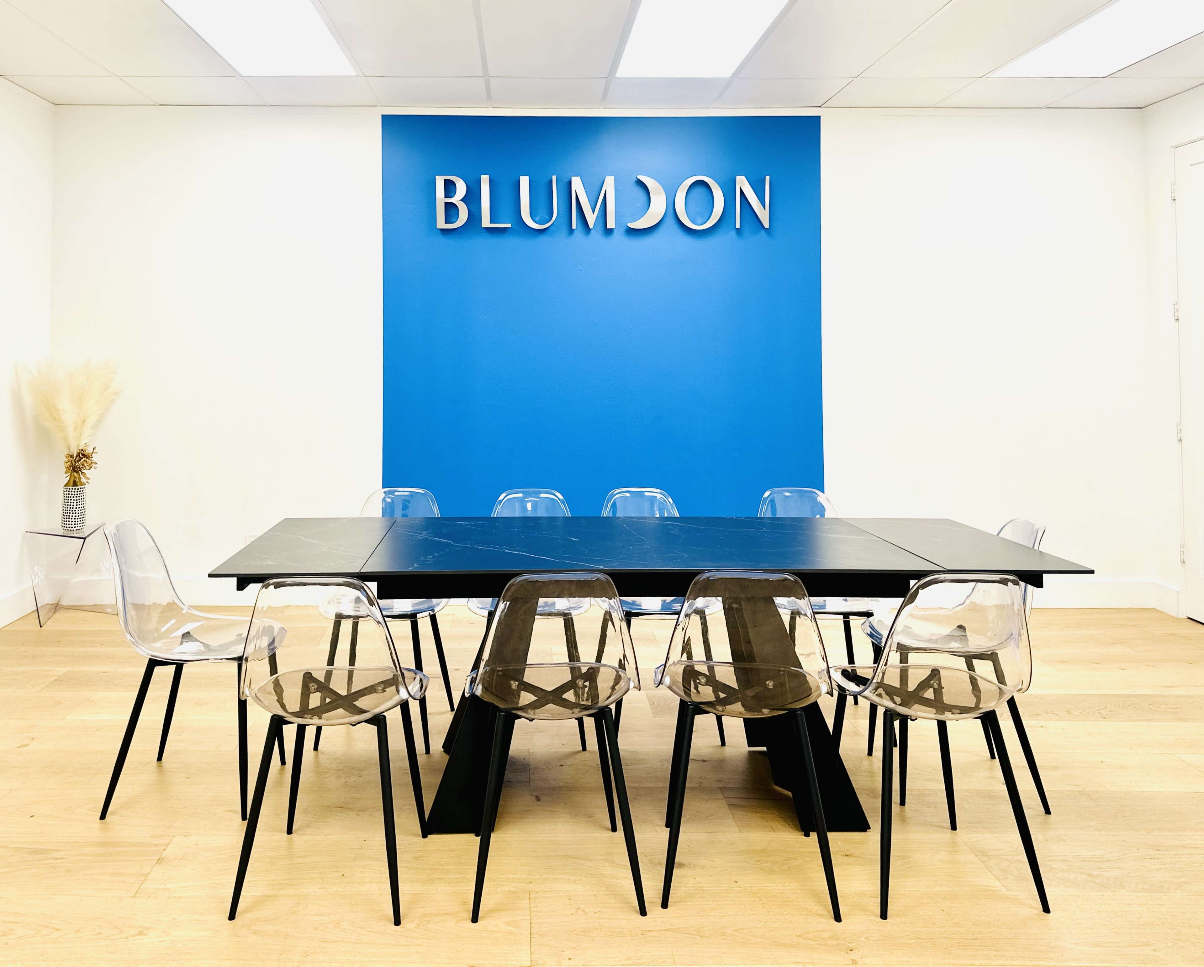A modern conference room features a long black table surrounded by transparent chairs, with a blue wall displaying the name "BLUMDON."