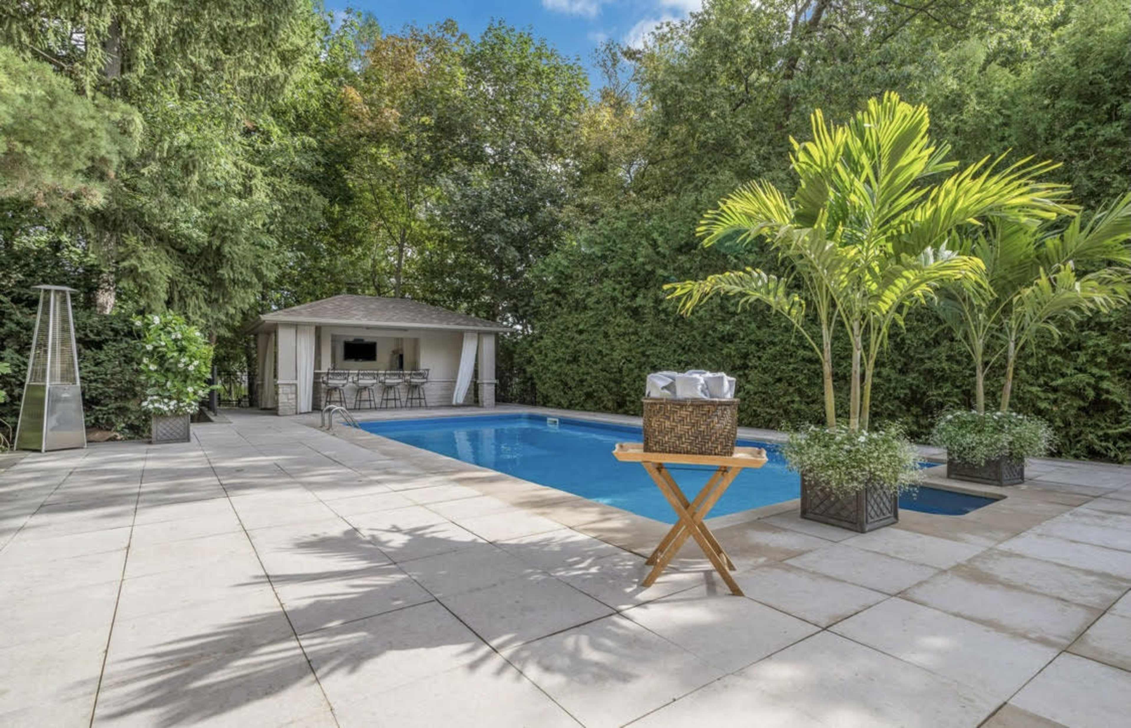 A rectangular swimming pool is surrounded by tiled flooring, lush greenery, and features a gazebo with seating in the background.