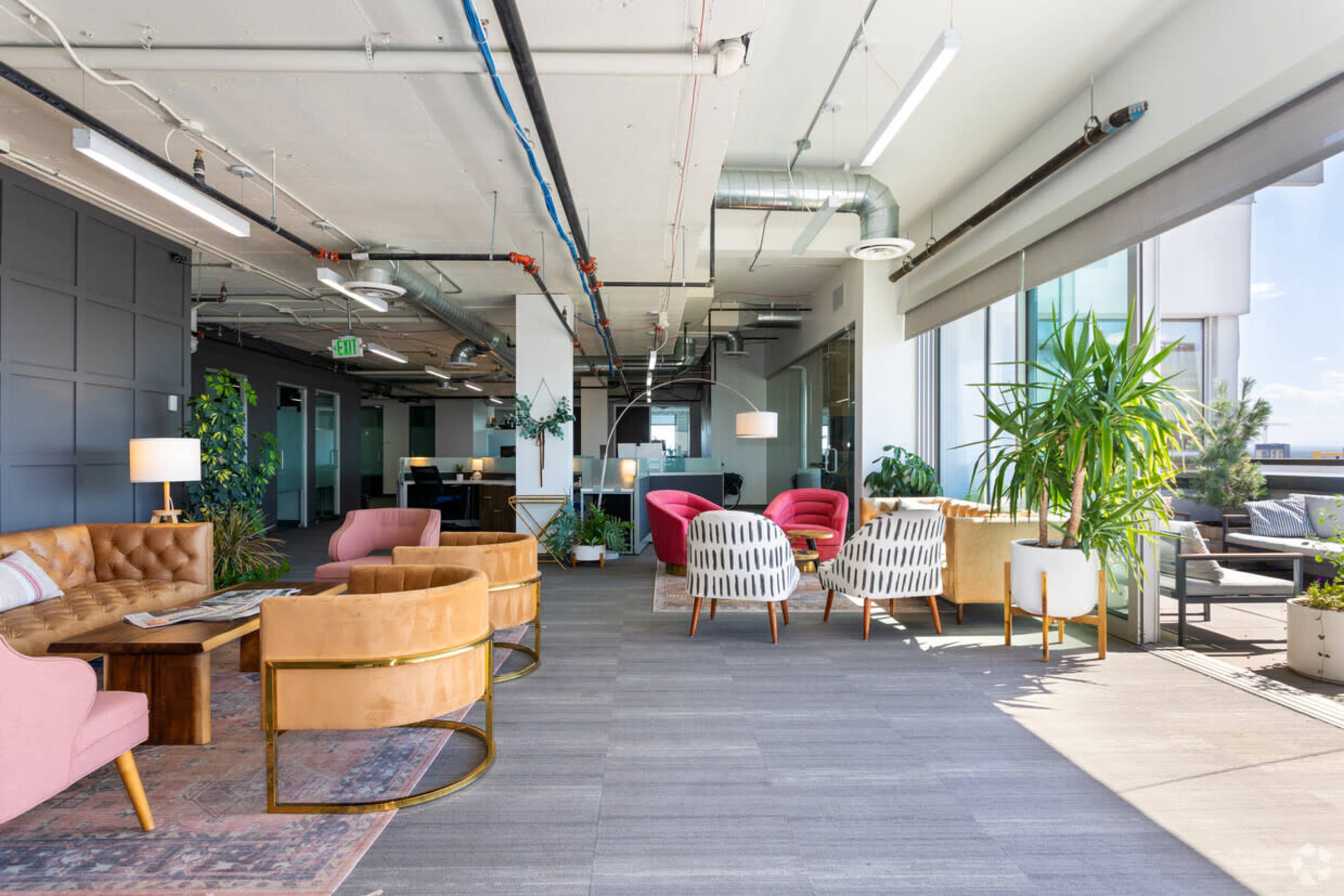 A modern office space featuring various seating areas with colorful furniture, large windows, and indoor plants.