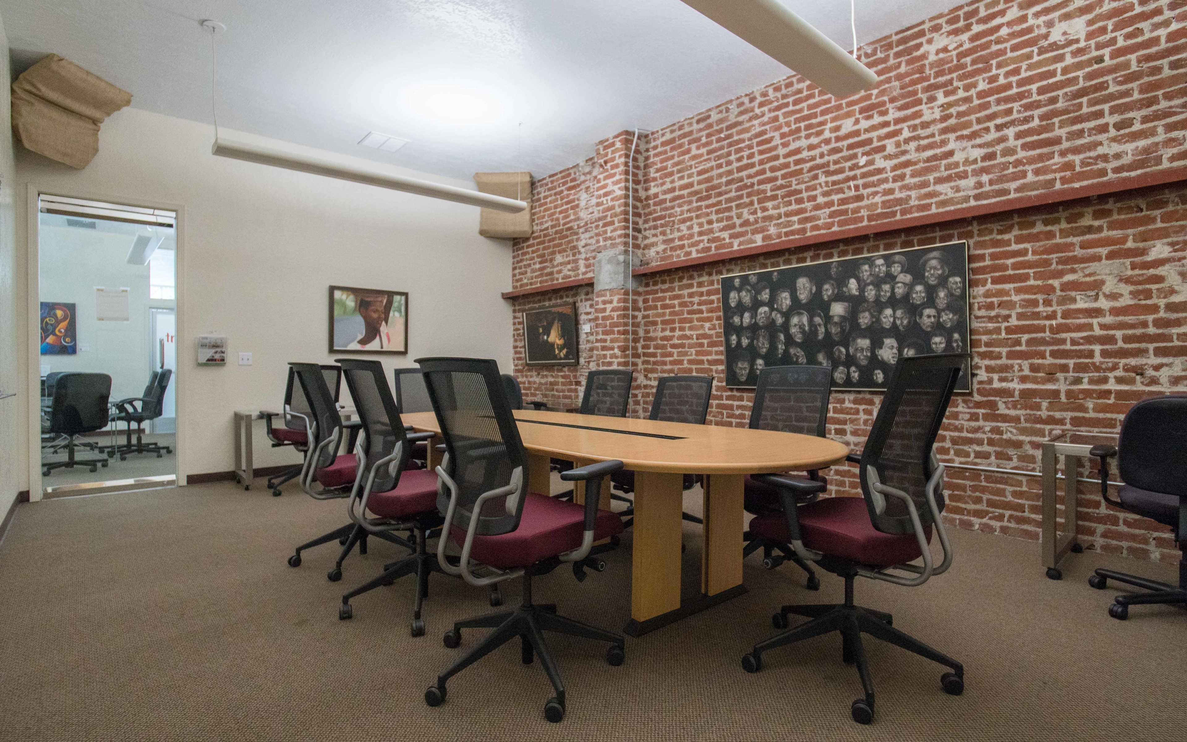 A conference room features a long wooden table surrounded by black office chairs, with exposed brick walls and framed artwork.