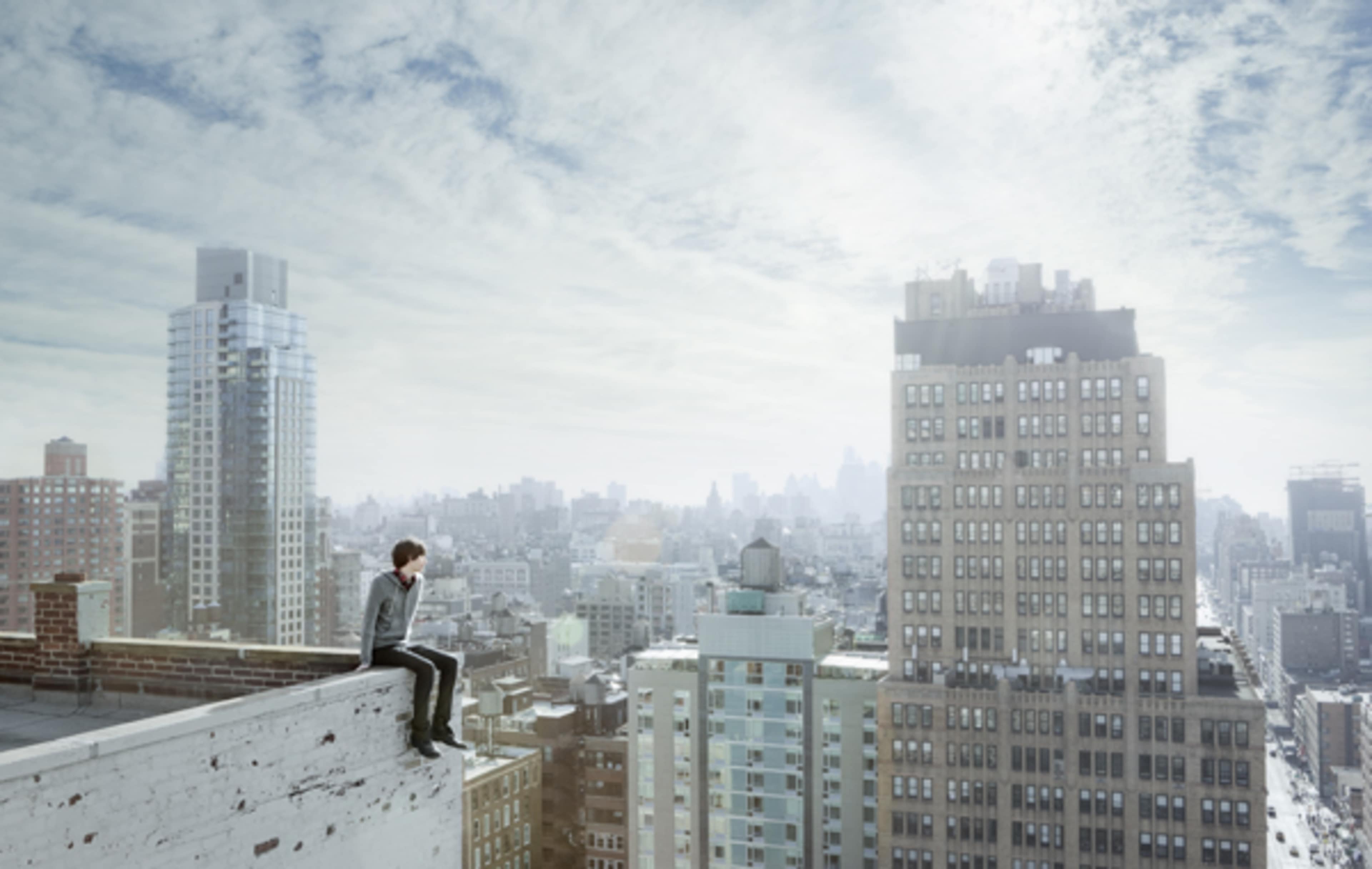 A person sits on the edge of a rooftop, overlooking a sprawling cityscape under a cloudy sky.