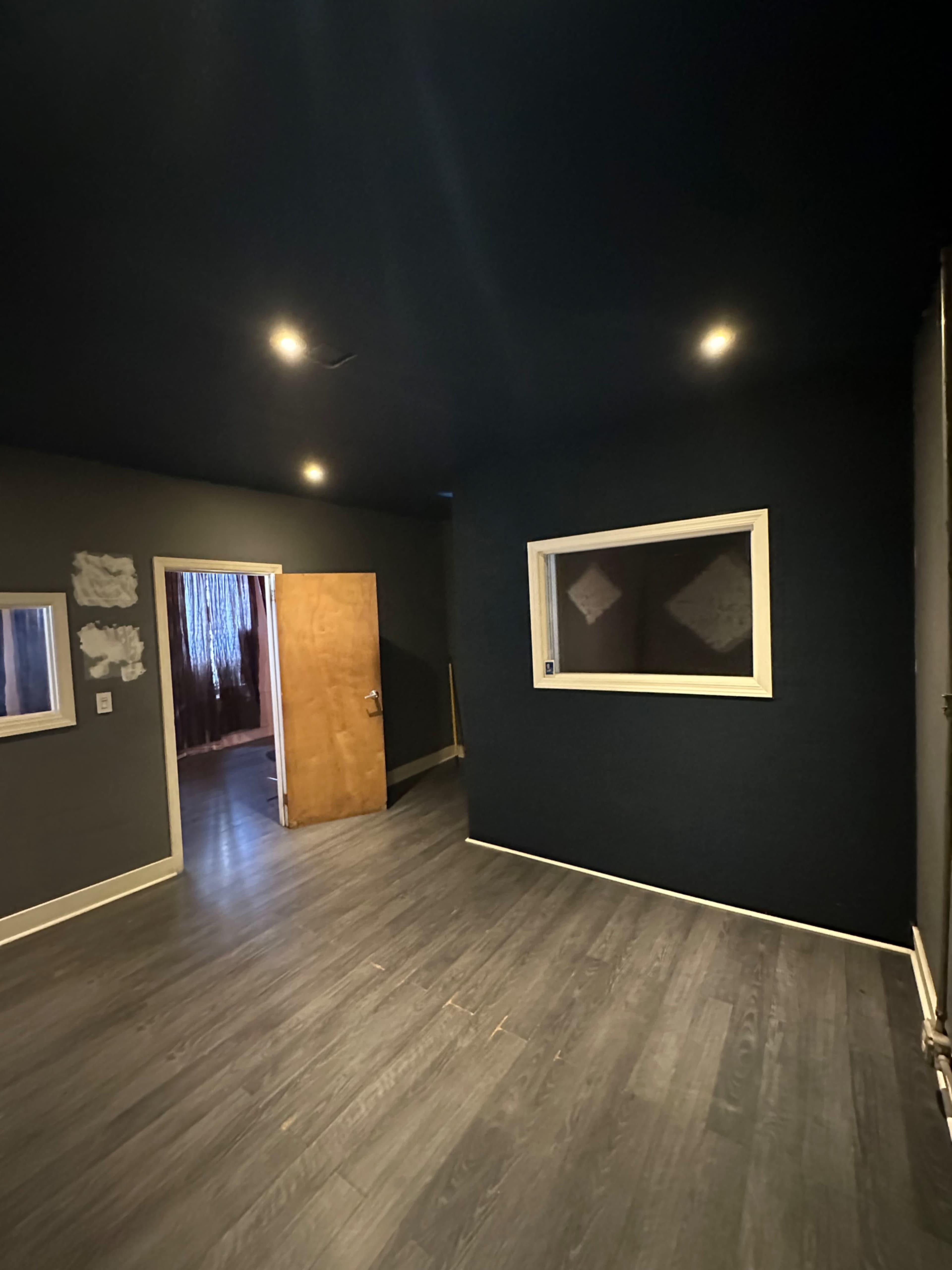 A dimly lit room with dark walls, a wooden door open to another space, and a large window on one wall.