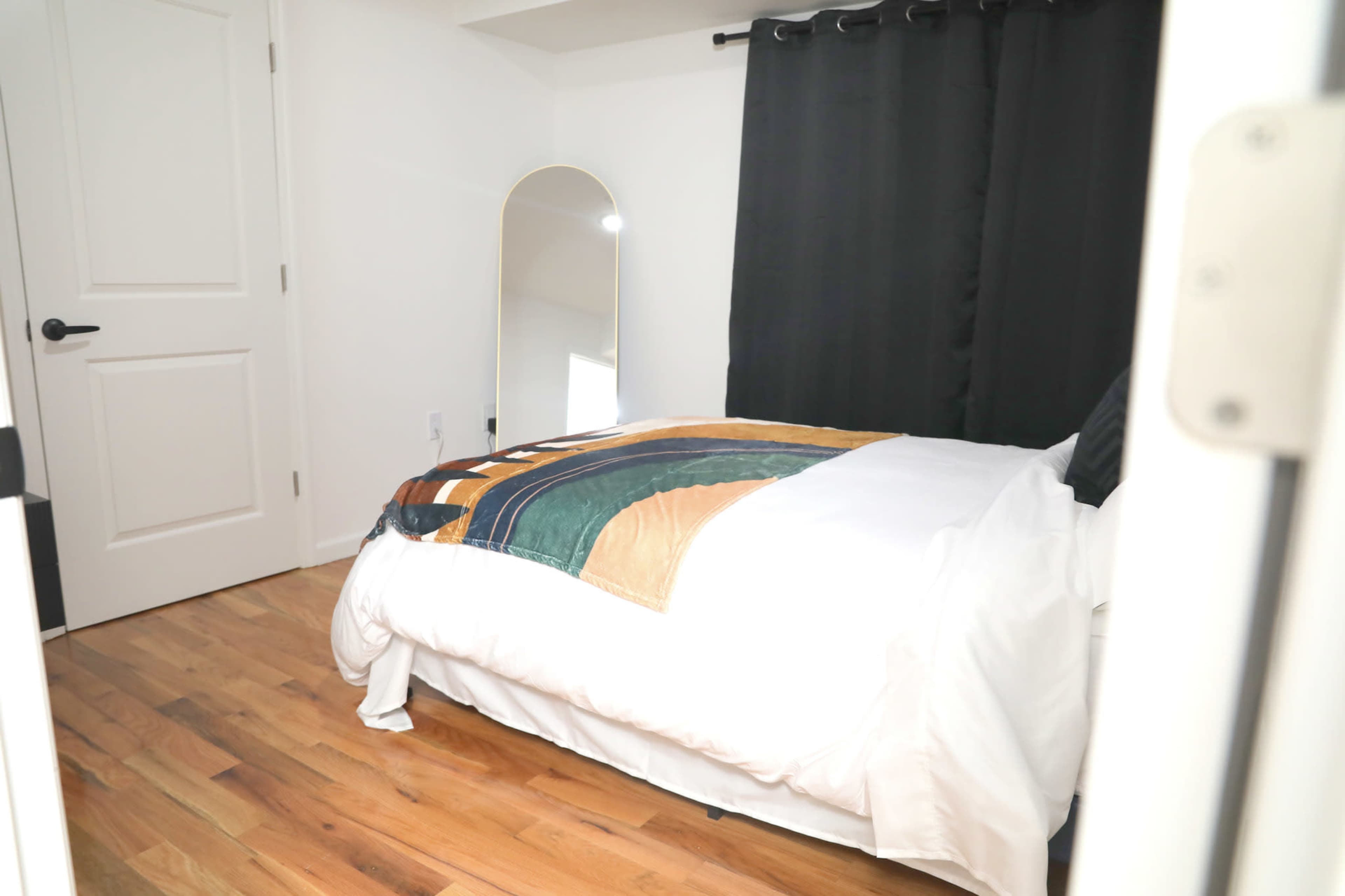 The image shows a bedroom featuring a bed with a patterned blanket, a full-length mirror, and black curtains covering a window.