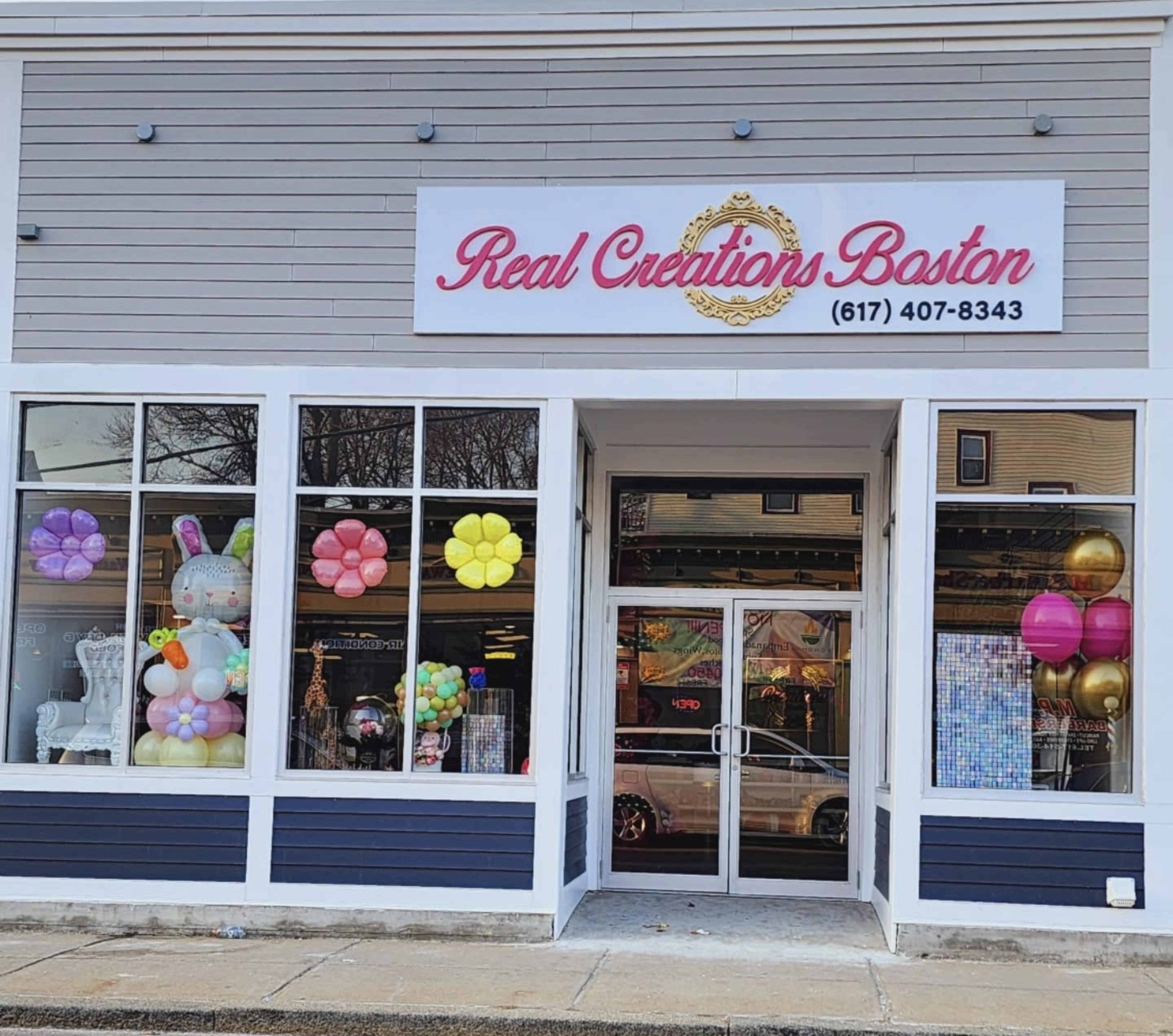 The storefront of Real Creations Boston features large windows decorated with colorful balloons and a prominent sign displaying the business name and contact number.