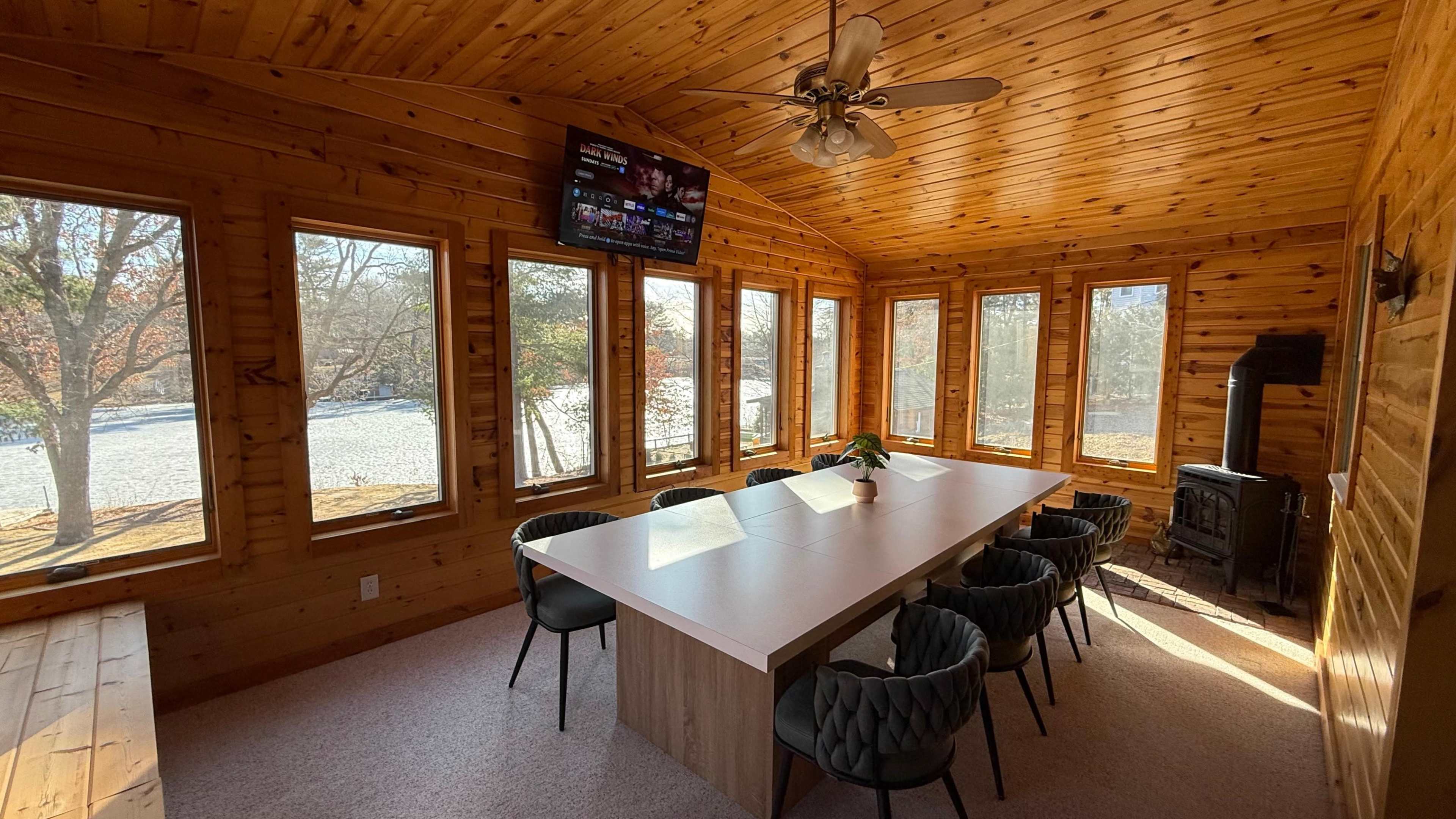 The image shows a wooden sunroom with large windows, featuring a long dining table surrounded by chairs and a television mounted on the wall.
