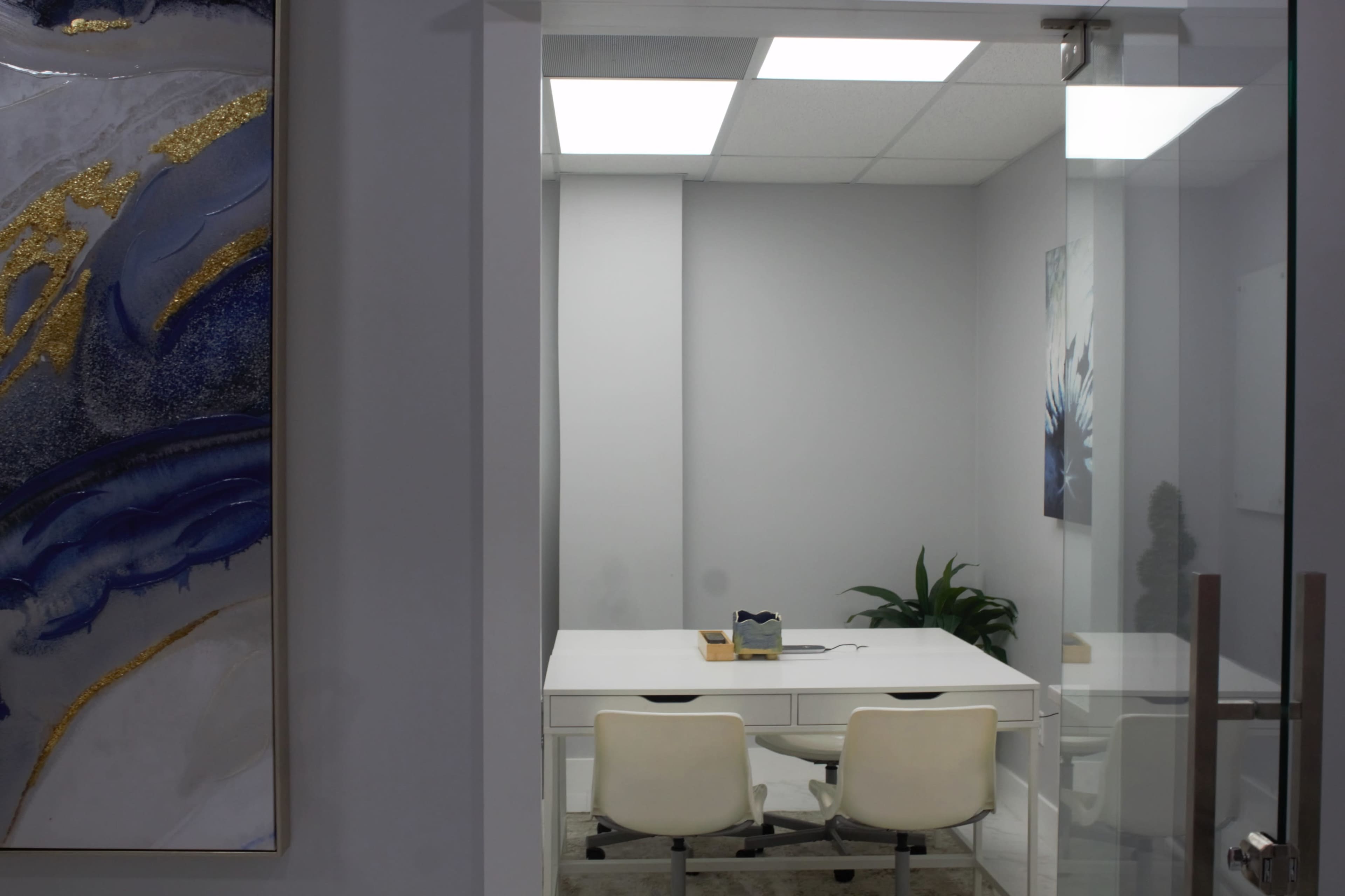 A small, modern office space features a white desk with two chairs, a plant, and a painting on the wall.