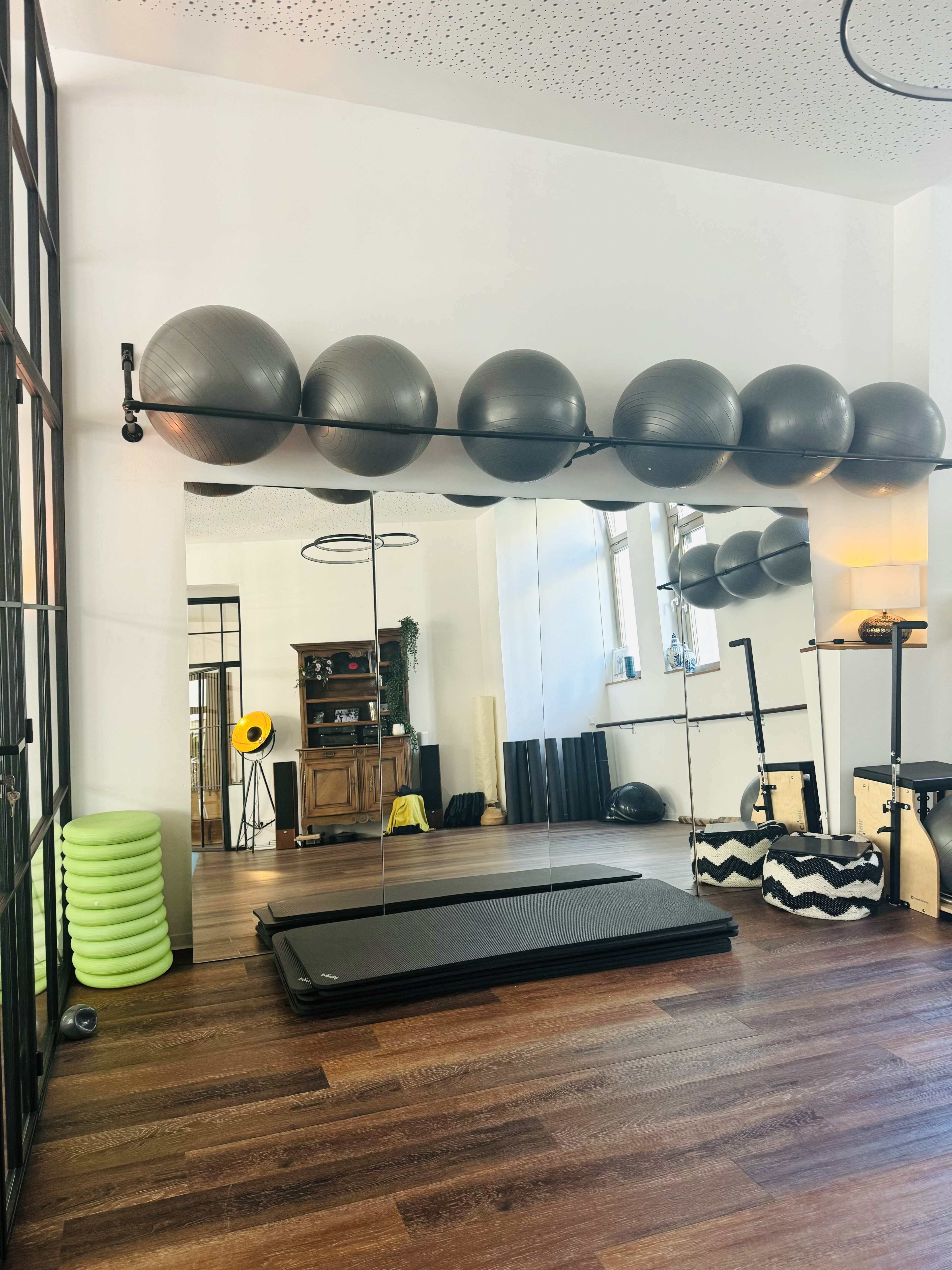 The image shows a well-lit exercise room featuring a wall mirror, exercise balls on a rack, and a variety of fitness equipment along the floor.