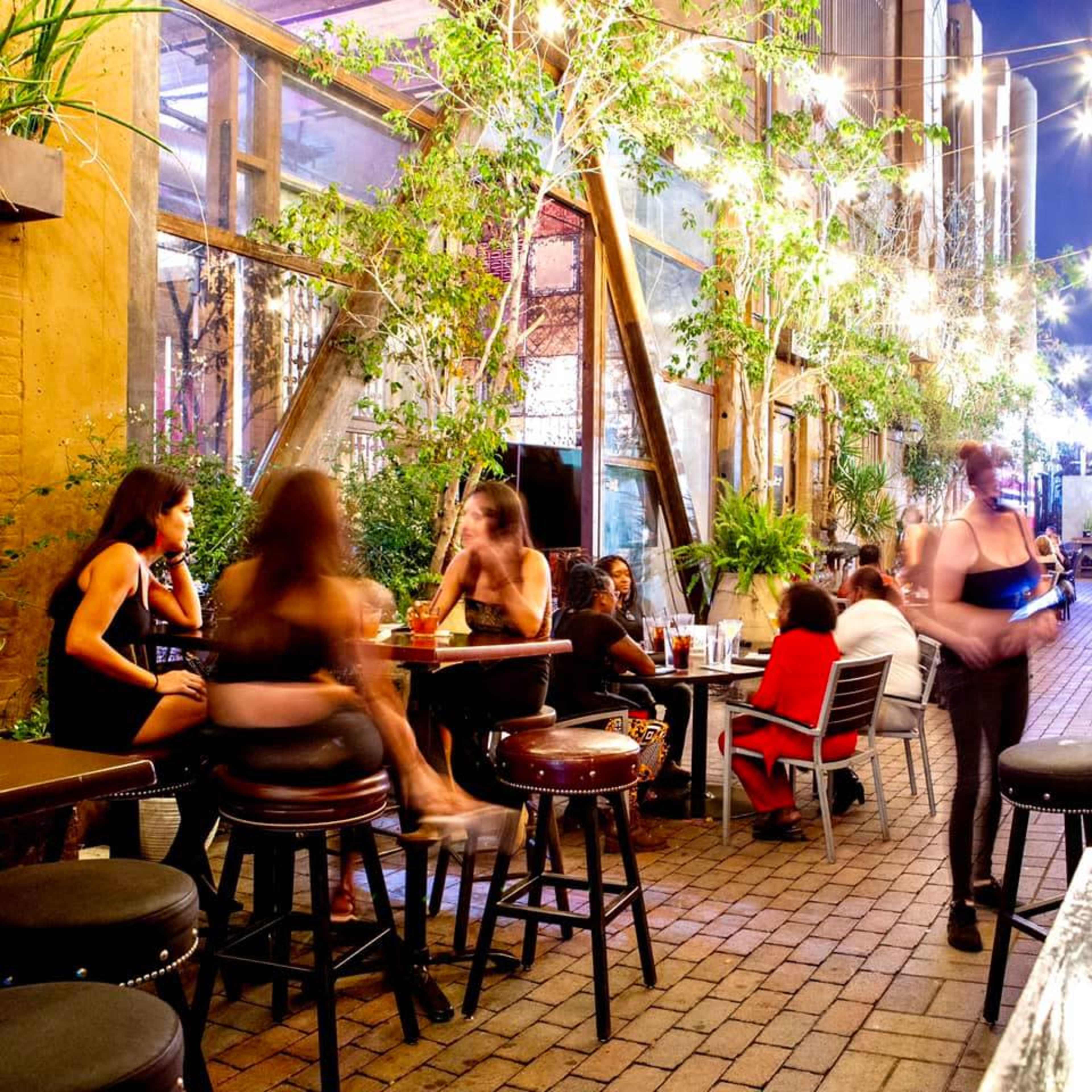 A lively outdoor dining area features patrons sitting at tables surrounded by plants and string lights in the evening.