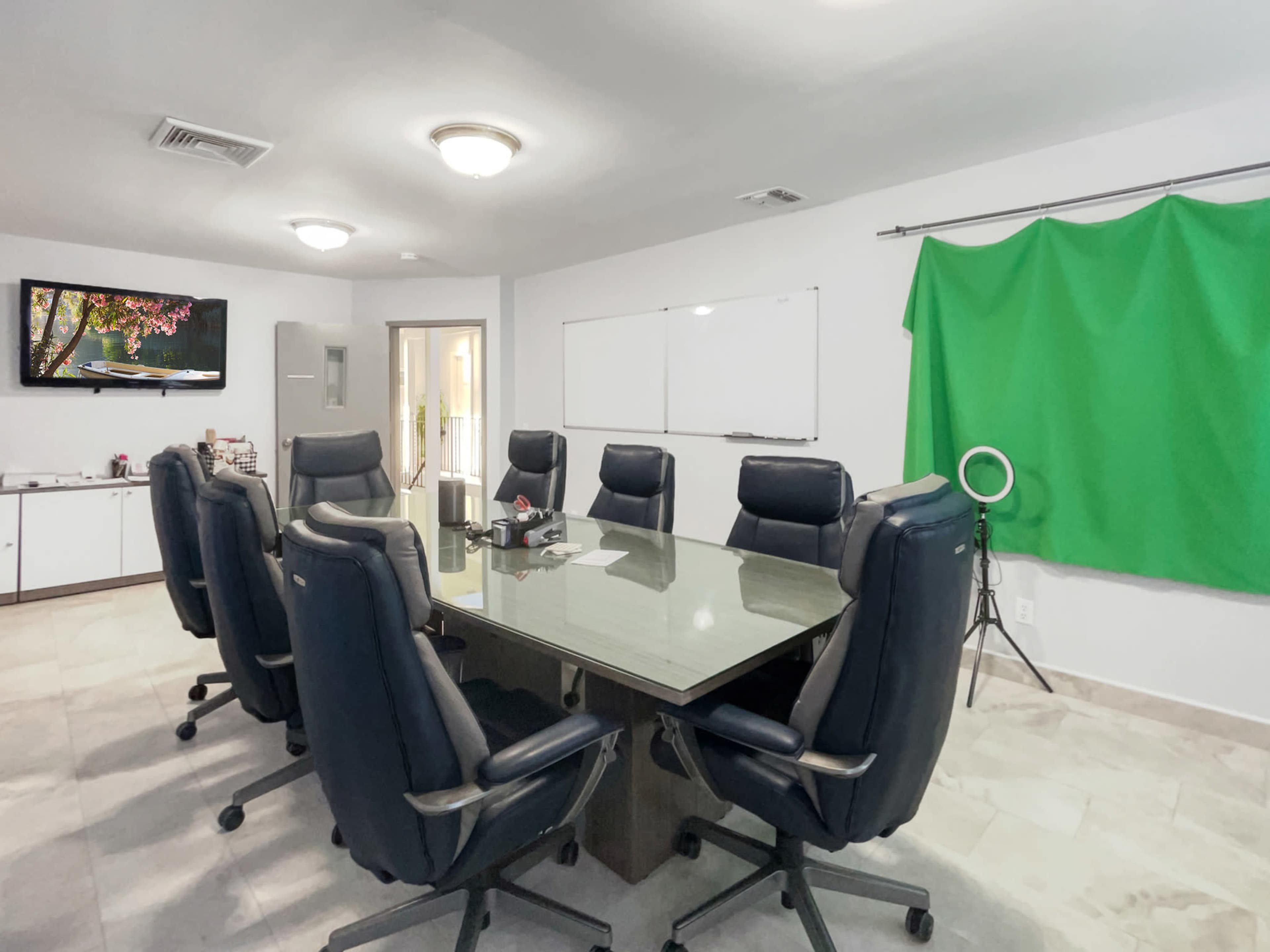 A conference room with a large rectangular table surrounded by eight black office chairs, a whiteboard on the wall, and a green backdrop on a stand.