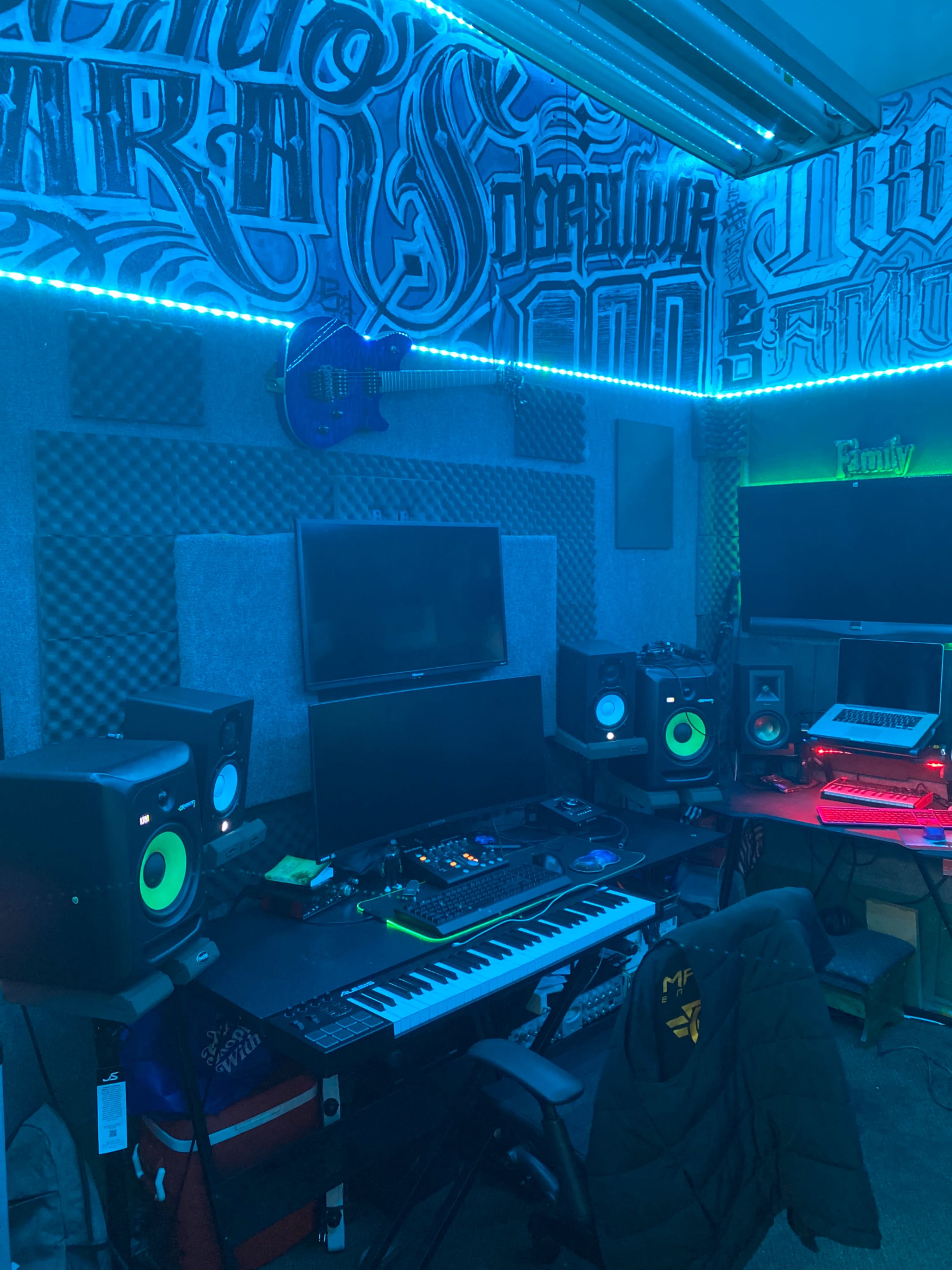 A music production studio with a computer monitor, keyboard, studio monitors, and colorful lighting against a graffiti-style wall.