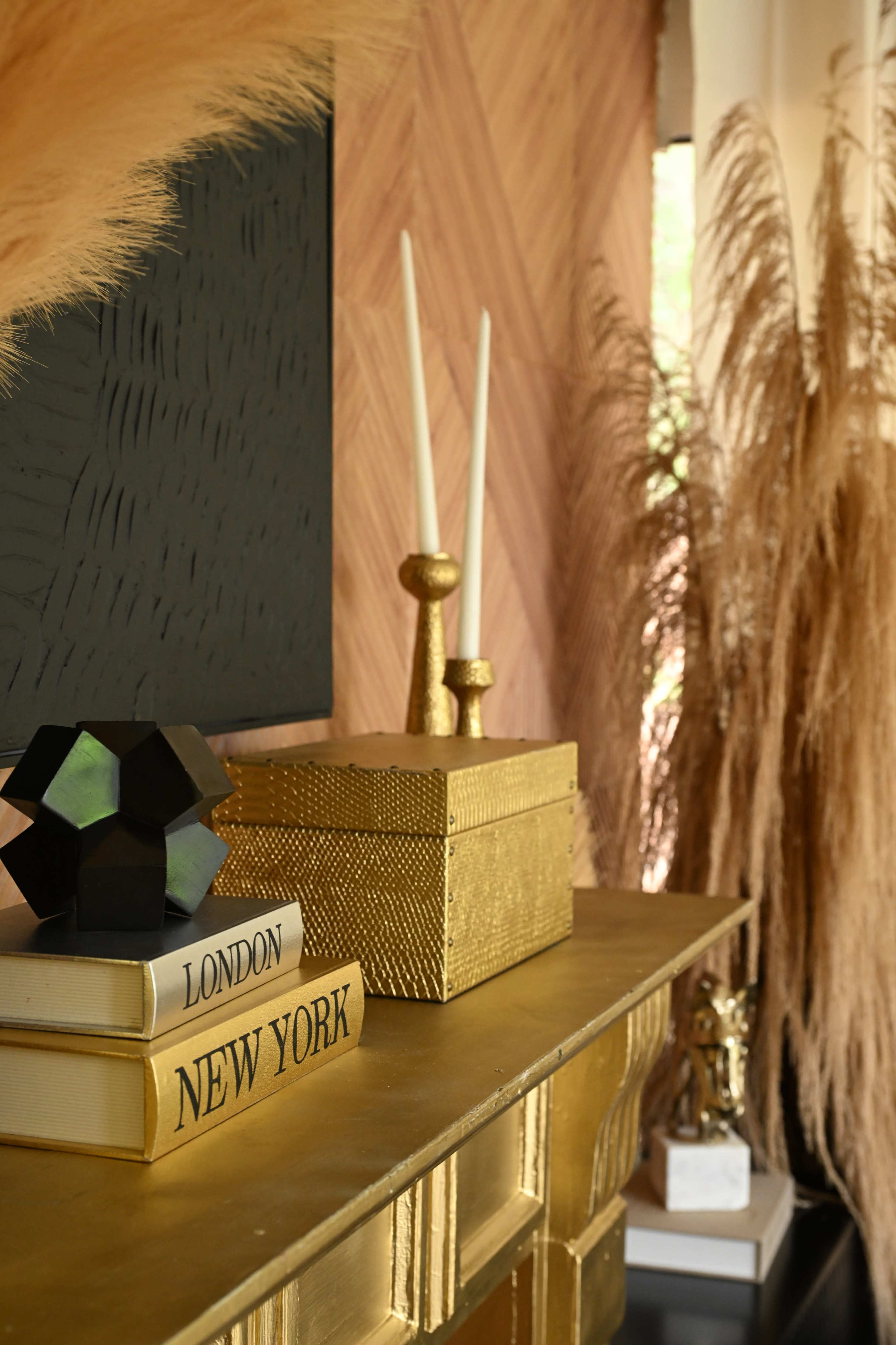 The image shows a decorative mantel with golden accents, featuring stacked books labeled "LONDON" and "NEW YORK," a geometric sculpture, and tall candlesticks beside pampas grass arrangements.