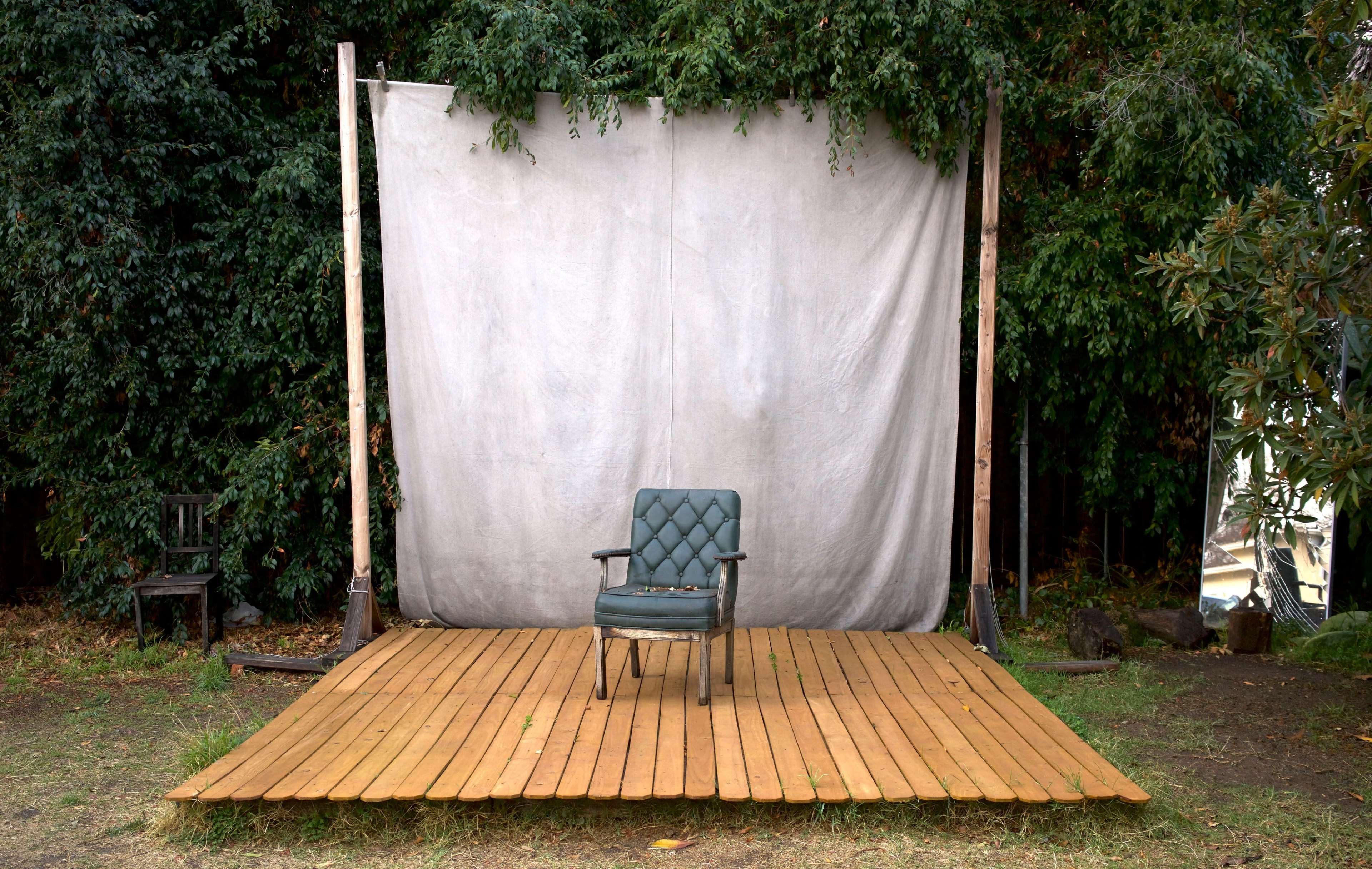 A green upholstered chair sits alone on a wooden platform in front of a white backdrop surrounded by greenery.