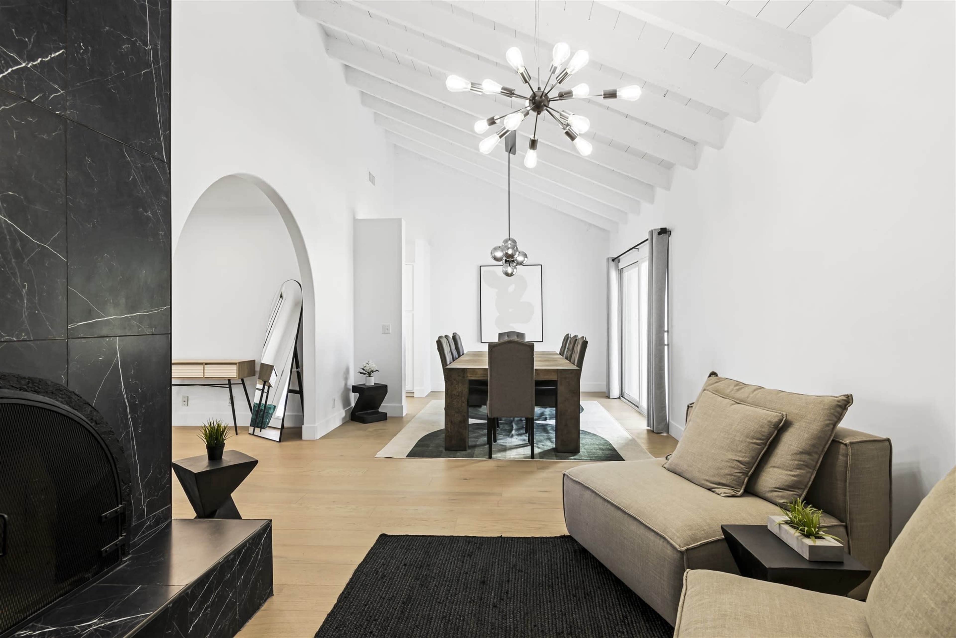 The image shows a modern, open-plan living space featuring a minimalist sofa, a dining area with a long table, and vaulted ceilings with a statement light fixture.