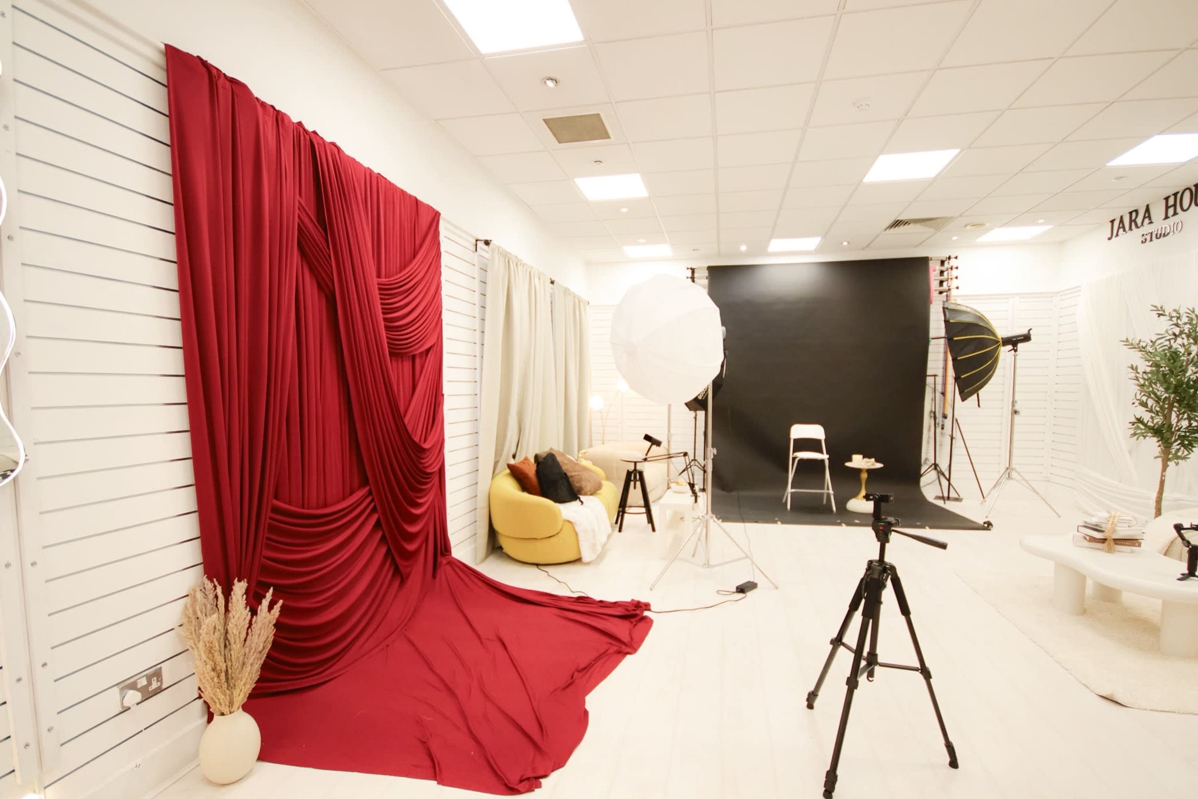 The image shows a photography studio featuring a red draped backdrop, a black backdrop setup, a light stand, and various props, including a yellow chair and a potted plant.