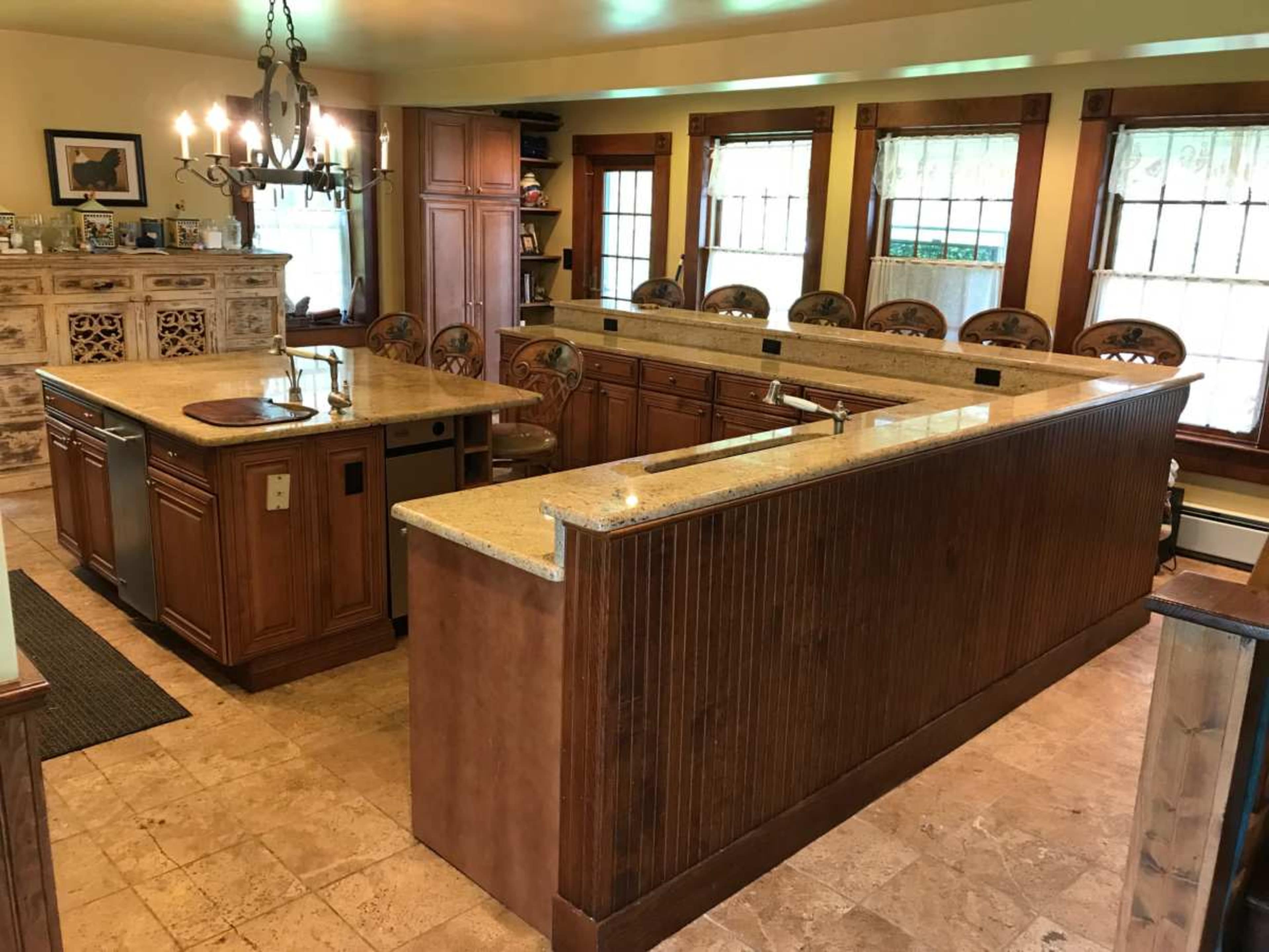The image shows a spacious kitchen with a large central island, wooden cabinetry, and several high-backed stools arranged around the island.