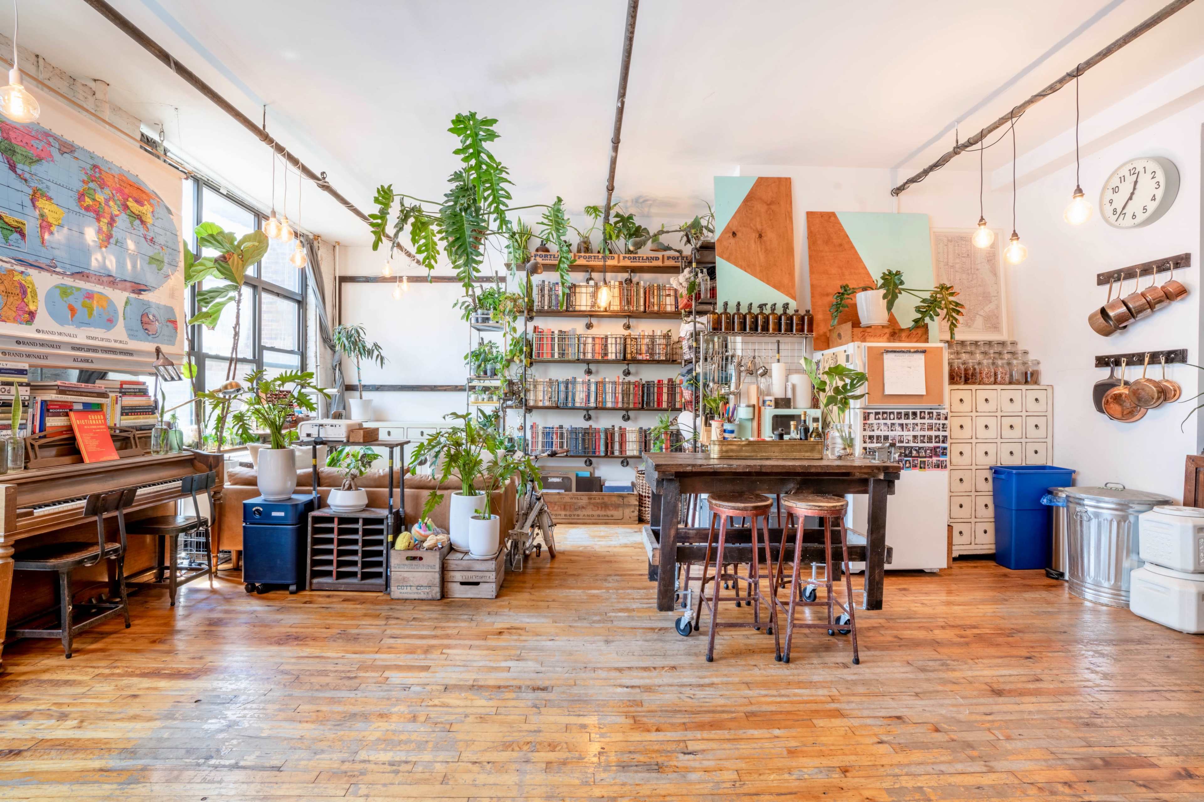 A bright, open space with wooden flooring, featuring a kitchen area with shelves lined with plants and various containers, a wooden table with bar stools, and a piano in the corner.