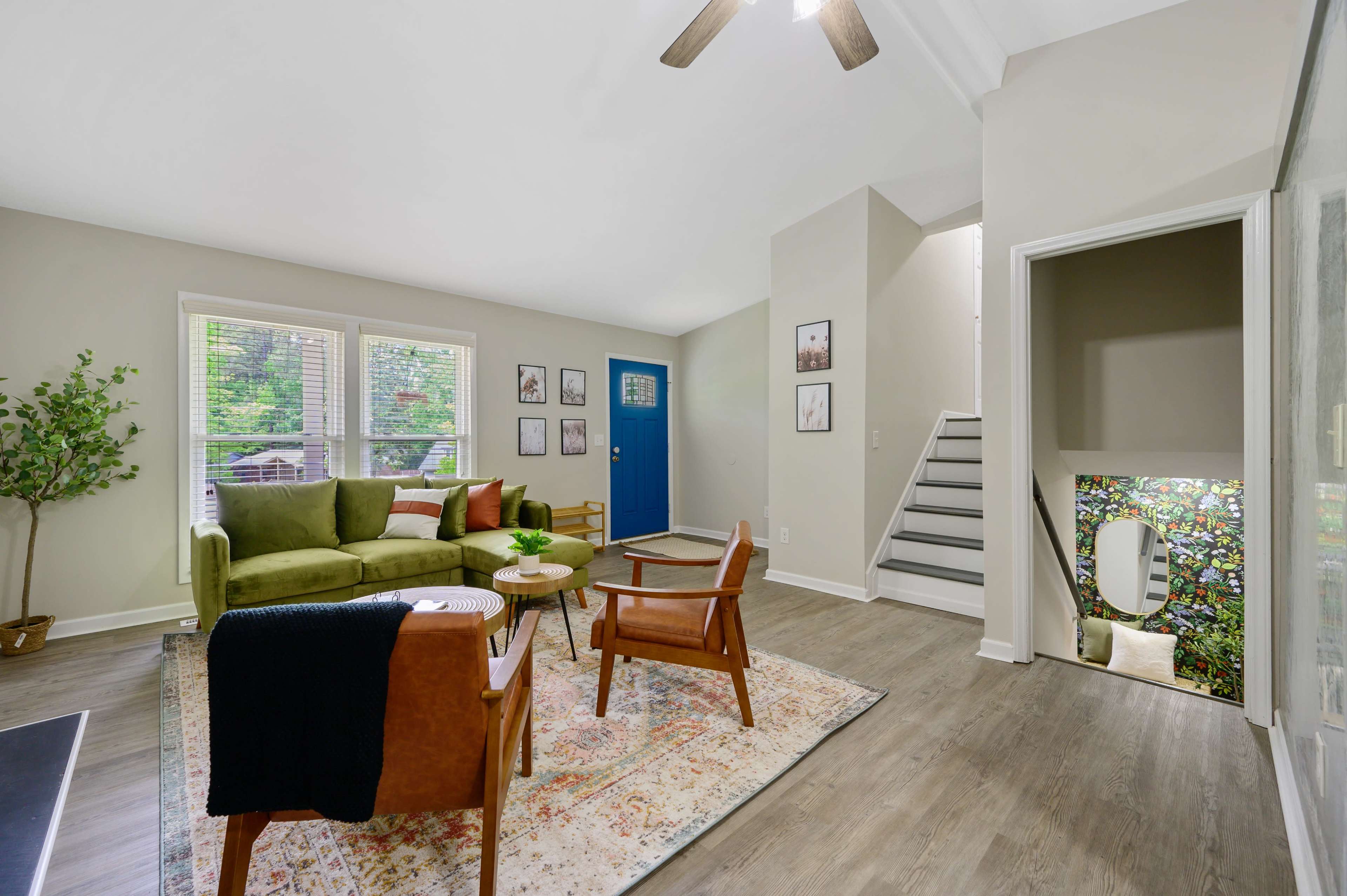 The image shows a living room with a green sofa, two wooden chairs, and a round coffee table, featuring a staircase and a colorful mirror in the corner.