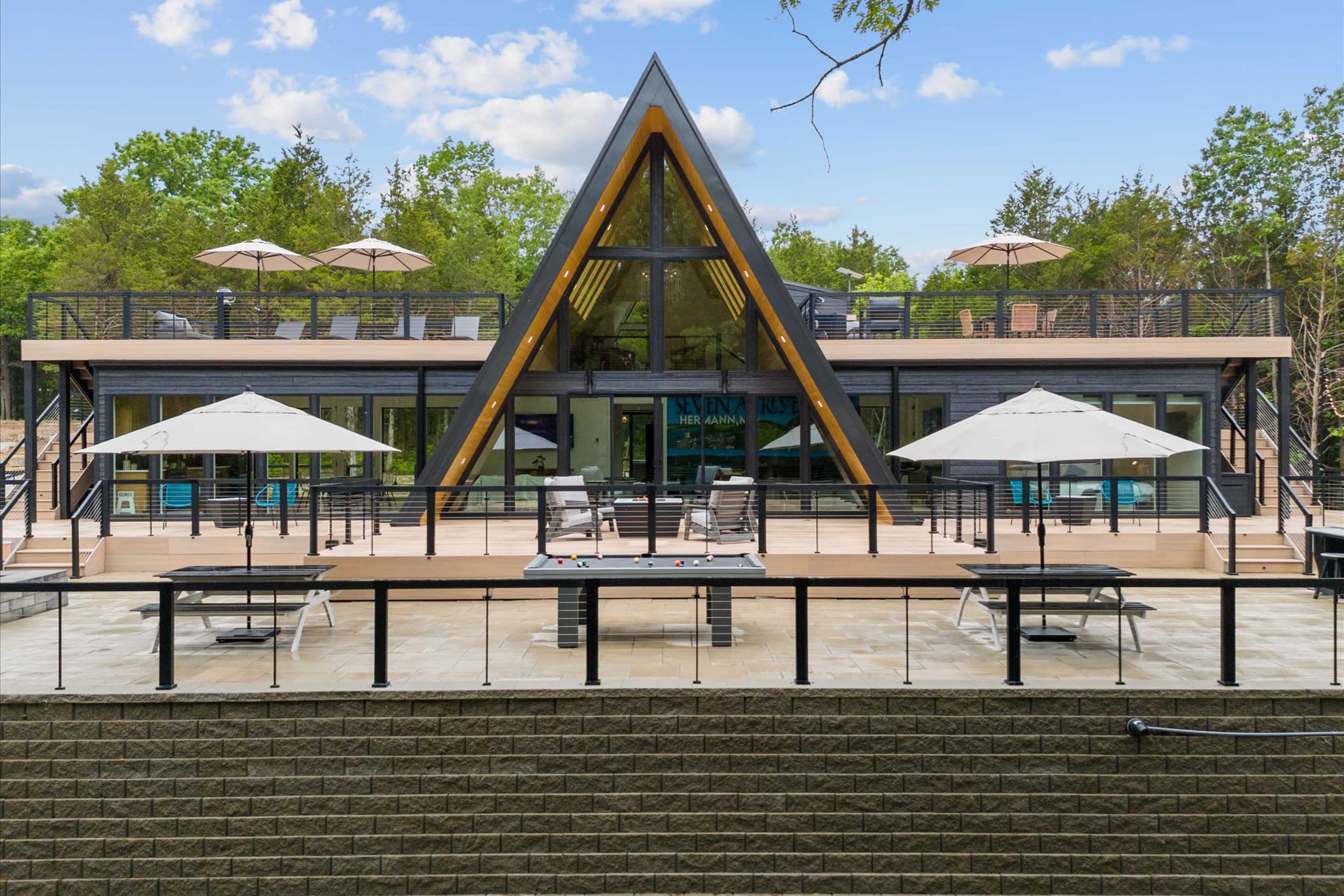 The image shows a modern A-frame house with large windows, a spacious deck featuring several lounge chairs and umbrellas, and a pool table on a patio area.