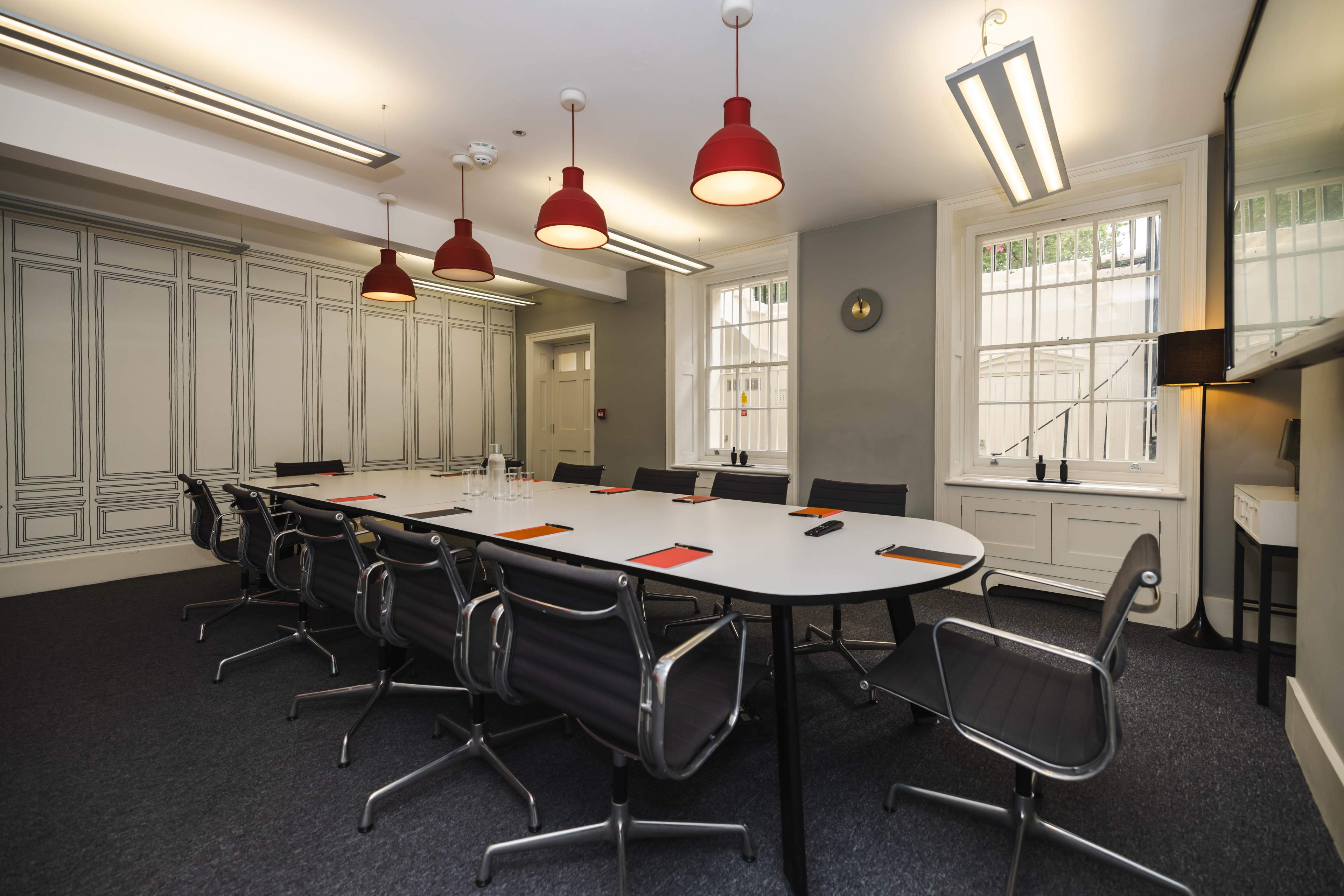 A modern conference room features a large oval table surrounded by black chairs, with red pendant lights overhead and large windows providing natural light.