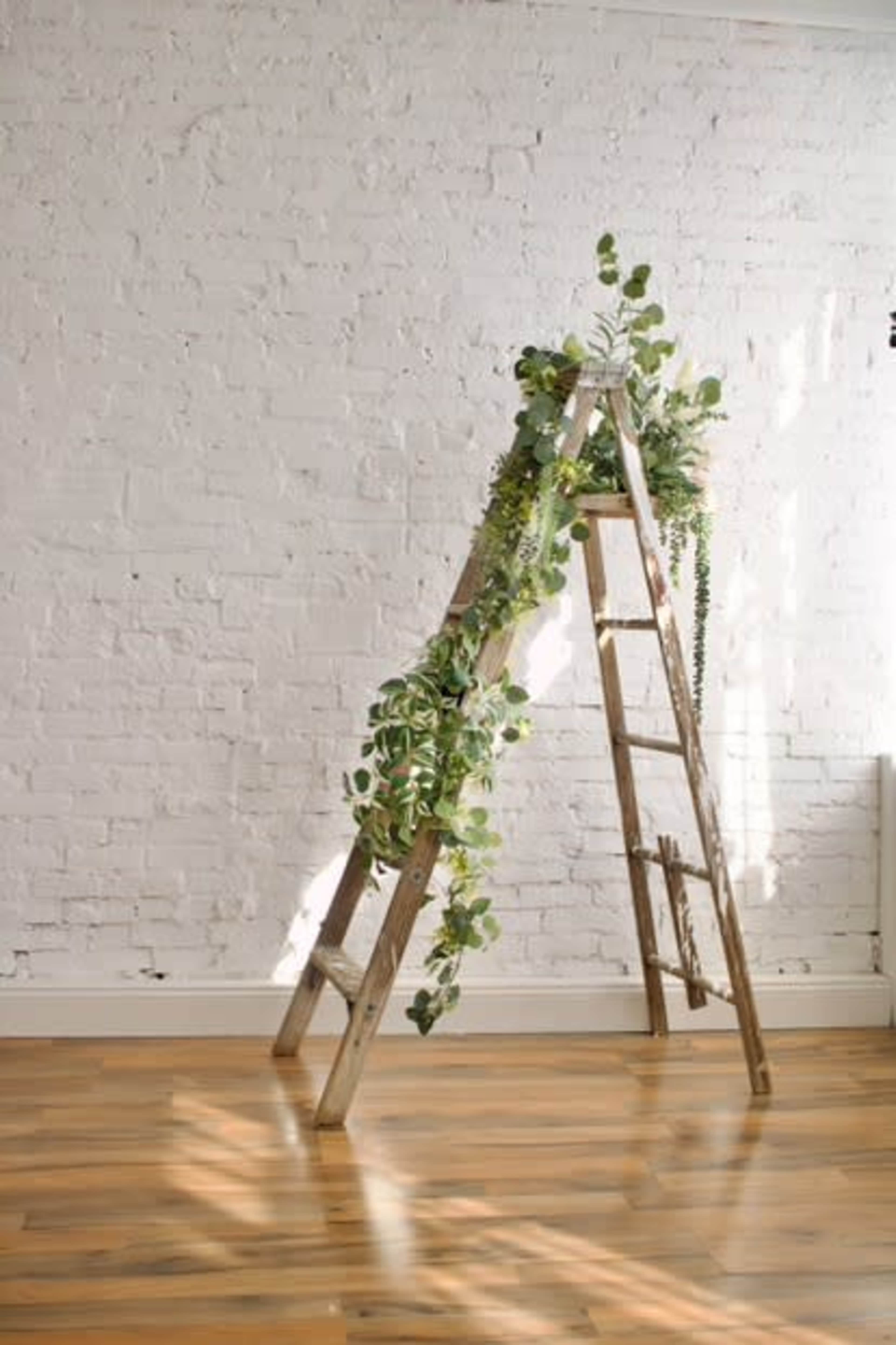 A wooden ladder is decorated with green plants against a white brick wall, placed on a wooden floor.