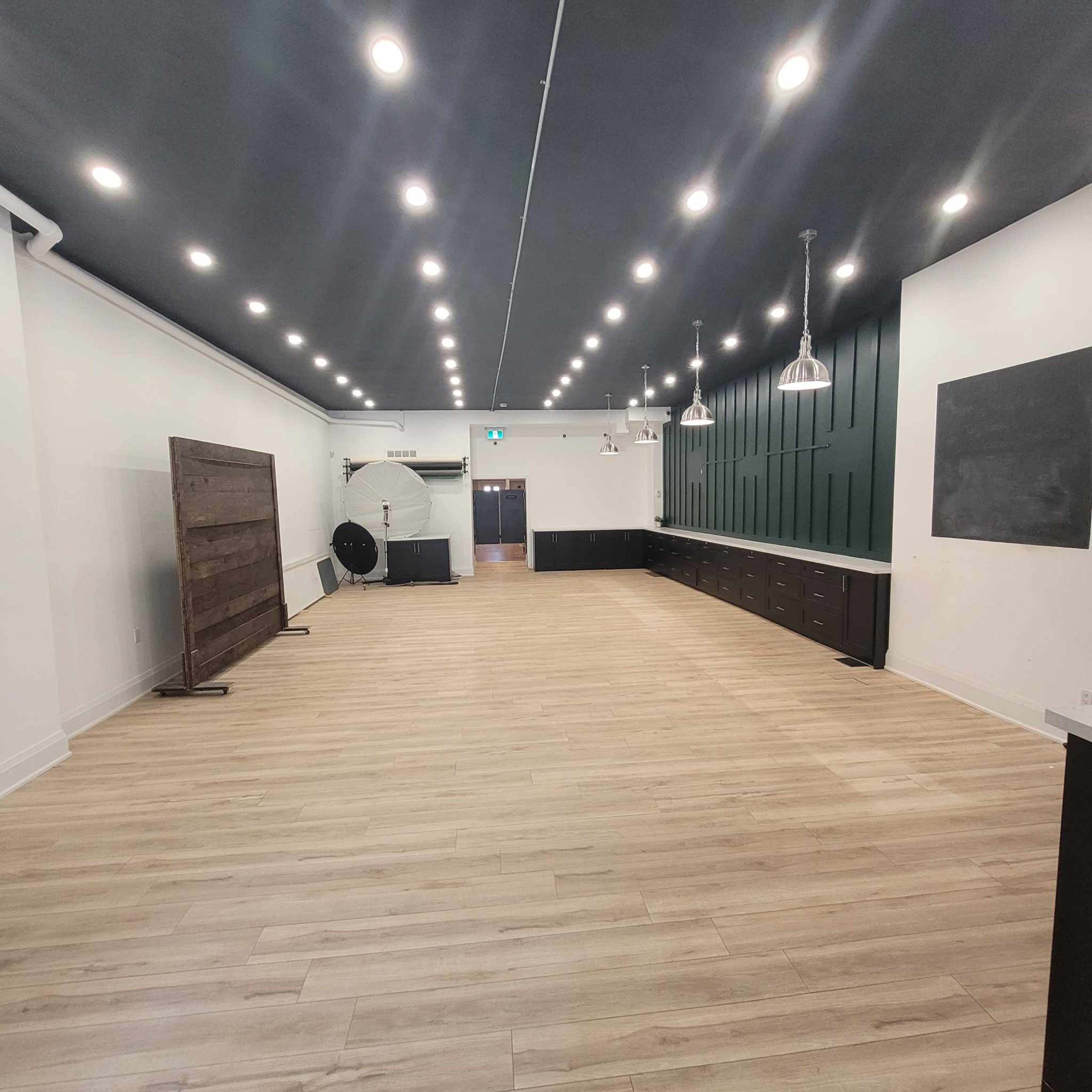 A spacious room with wooden flooring, high ceilings, and a combination of white and dark green walls, featuring multiple lights and a long counter along one side.