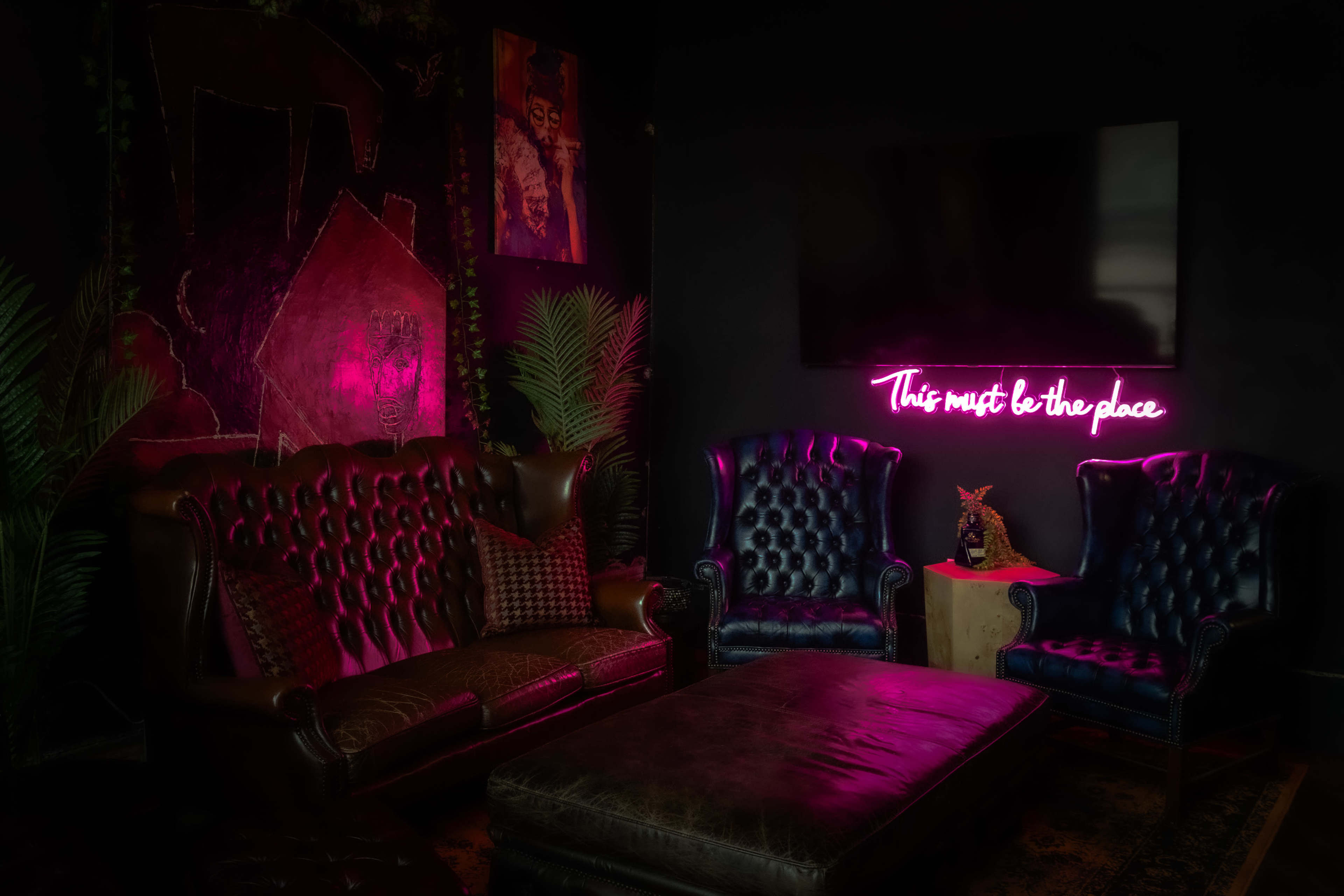 A cozy lounge area features two leather sofas, a low coffee table, and a neon sign that reads "This must be the place" against a dark wall adorned with art.