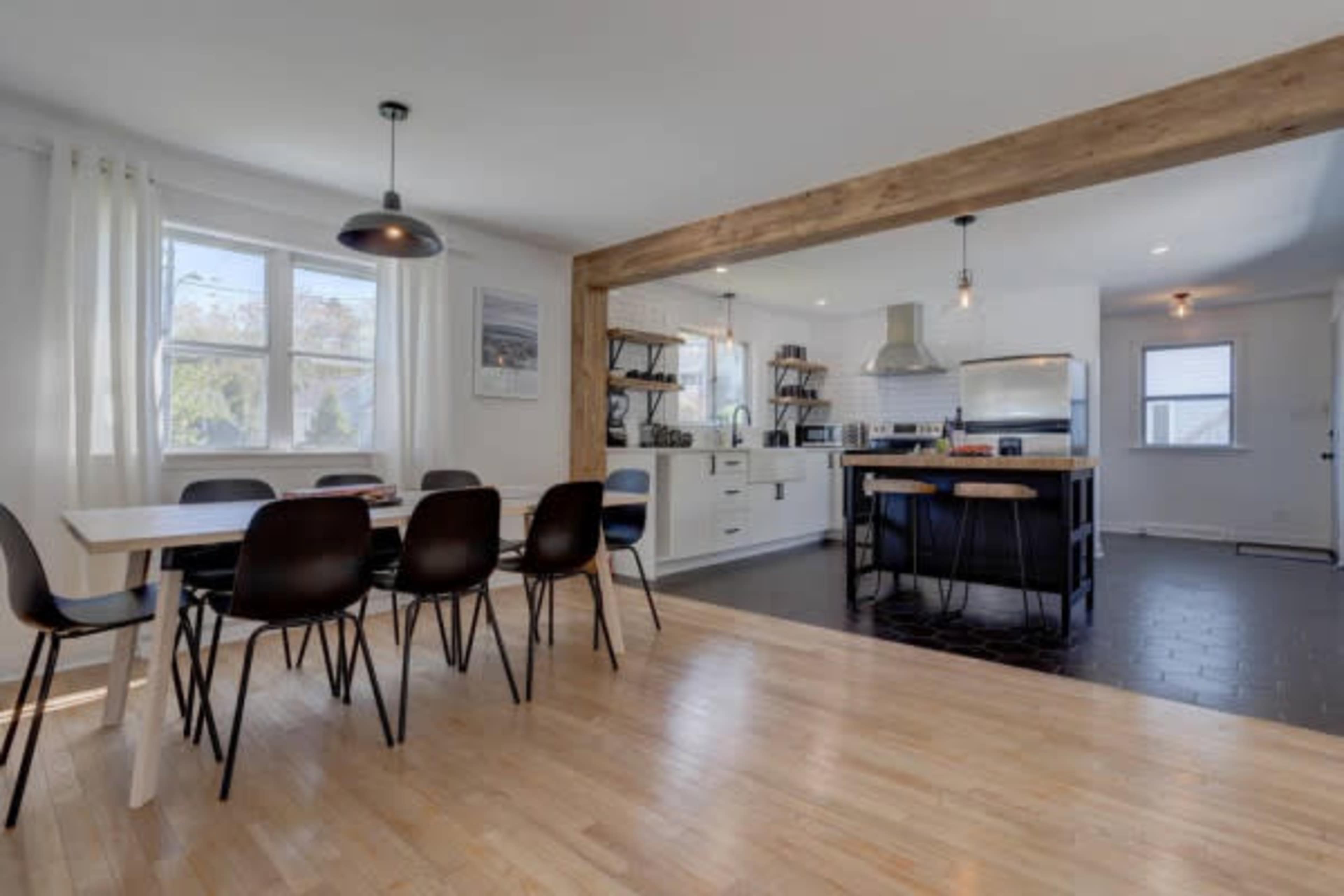 A modern kitchen and dining area feature a wooden beam, a dining table with chairs, and open shelving with kitchen appliances.