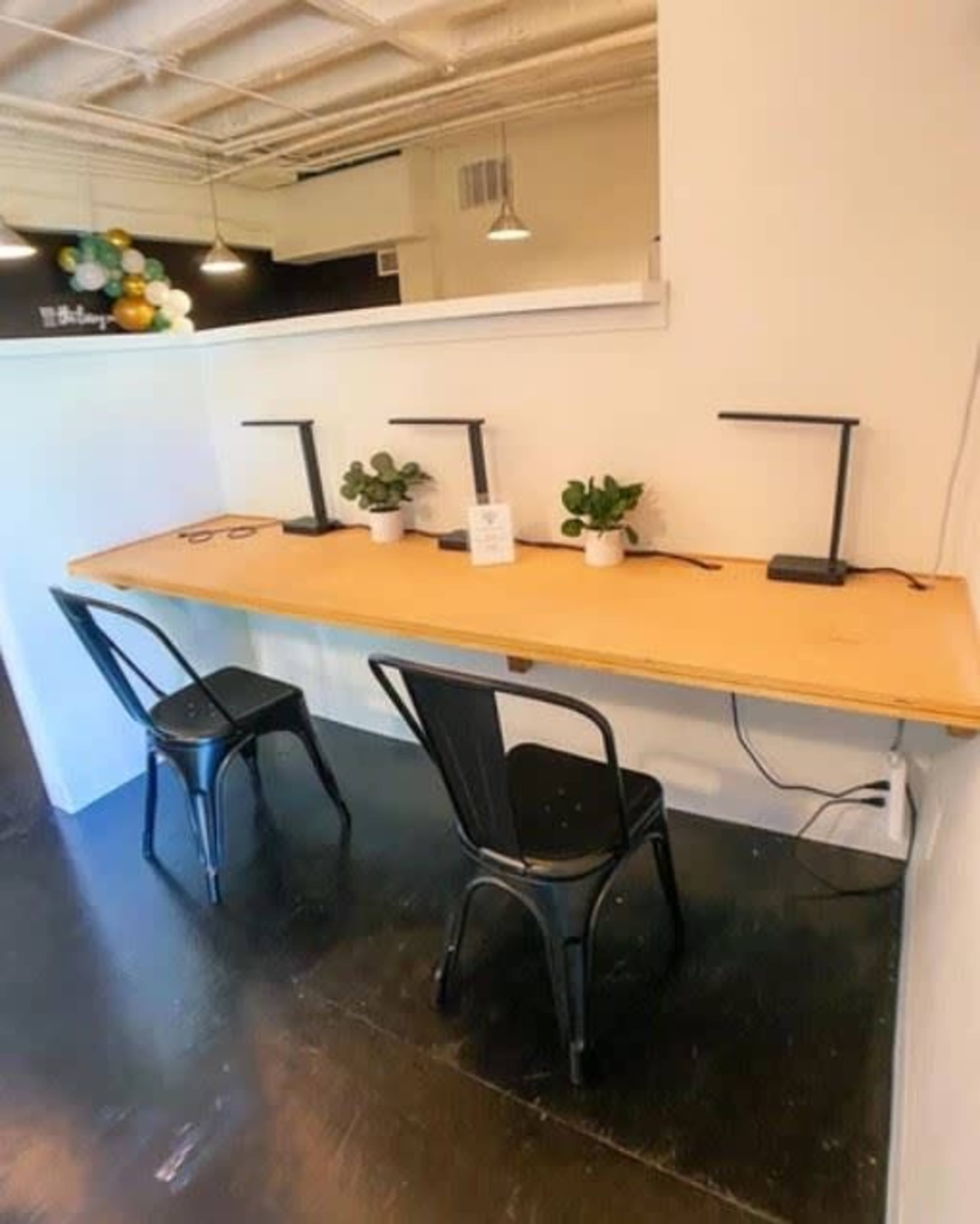 A room with a long wooden table featuring four desk lamps and two black chairs, along with small potted plants on the table.