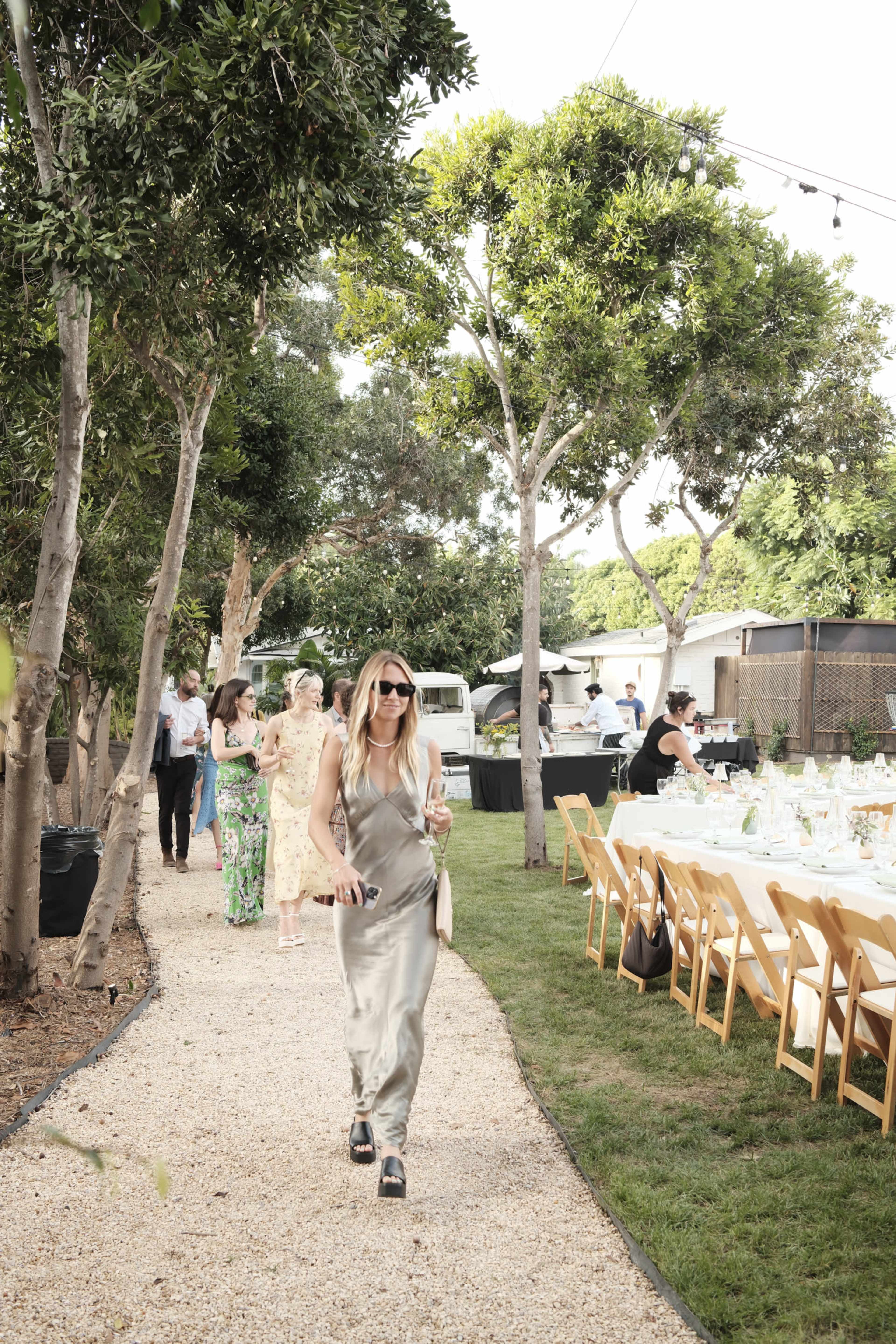 A woman in a silver dress walks along a gravel path in a garden setting with tables arranged for an outdoor event.