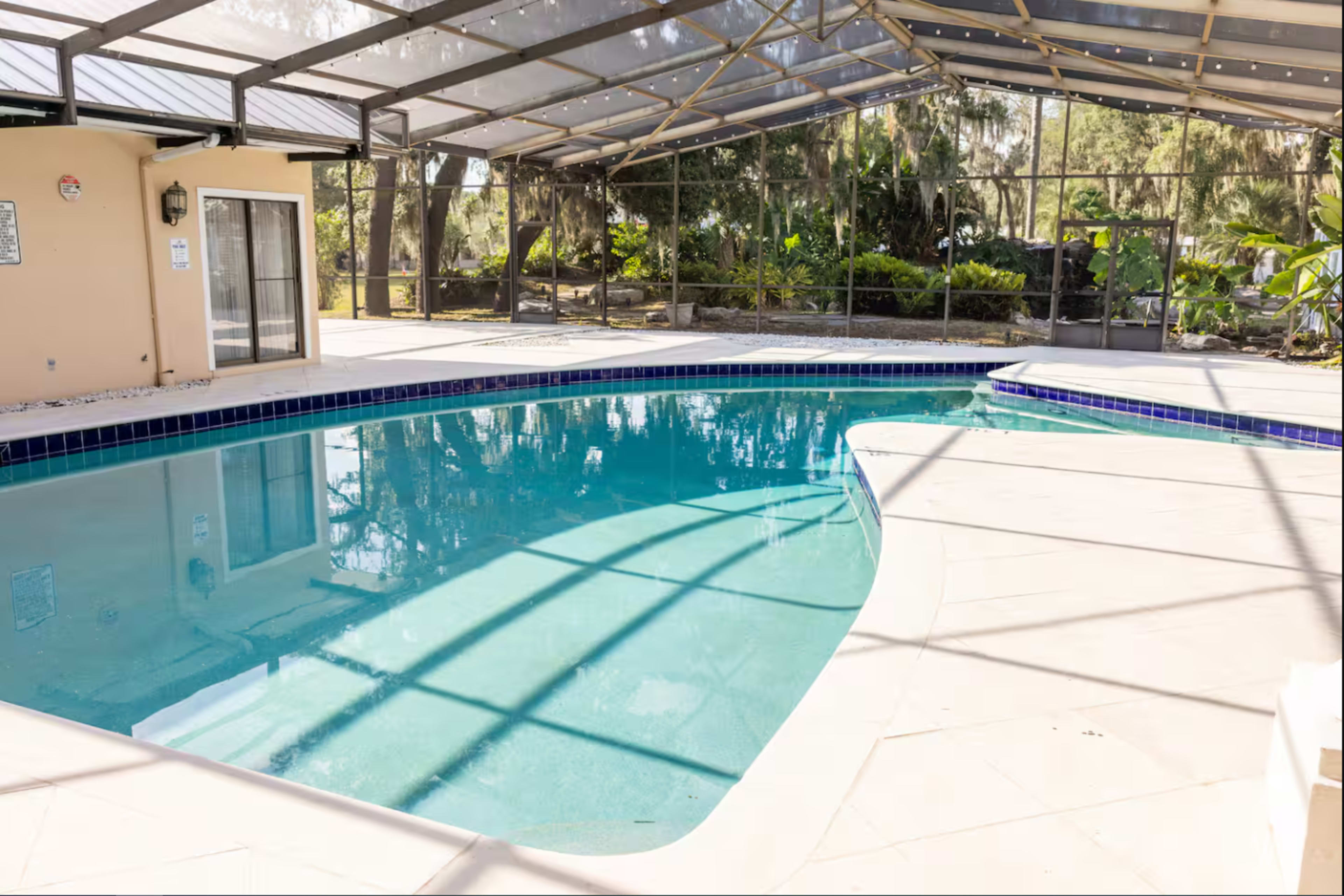 The image shows a clear blue swimming pool with a screened enclosure, surrounded by greenery and sunlight.