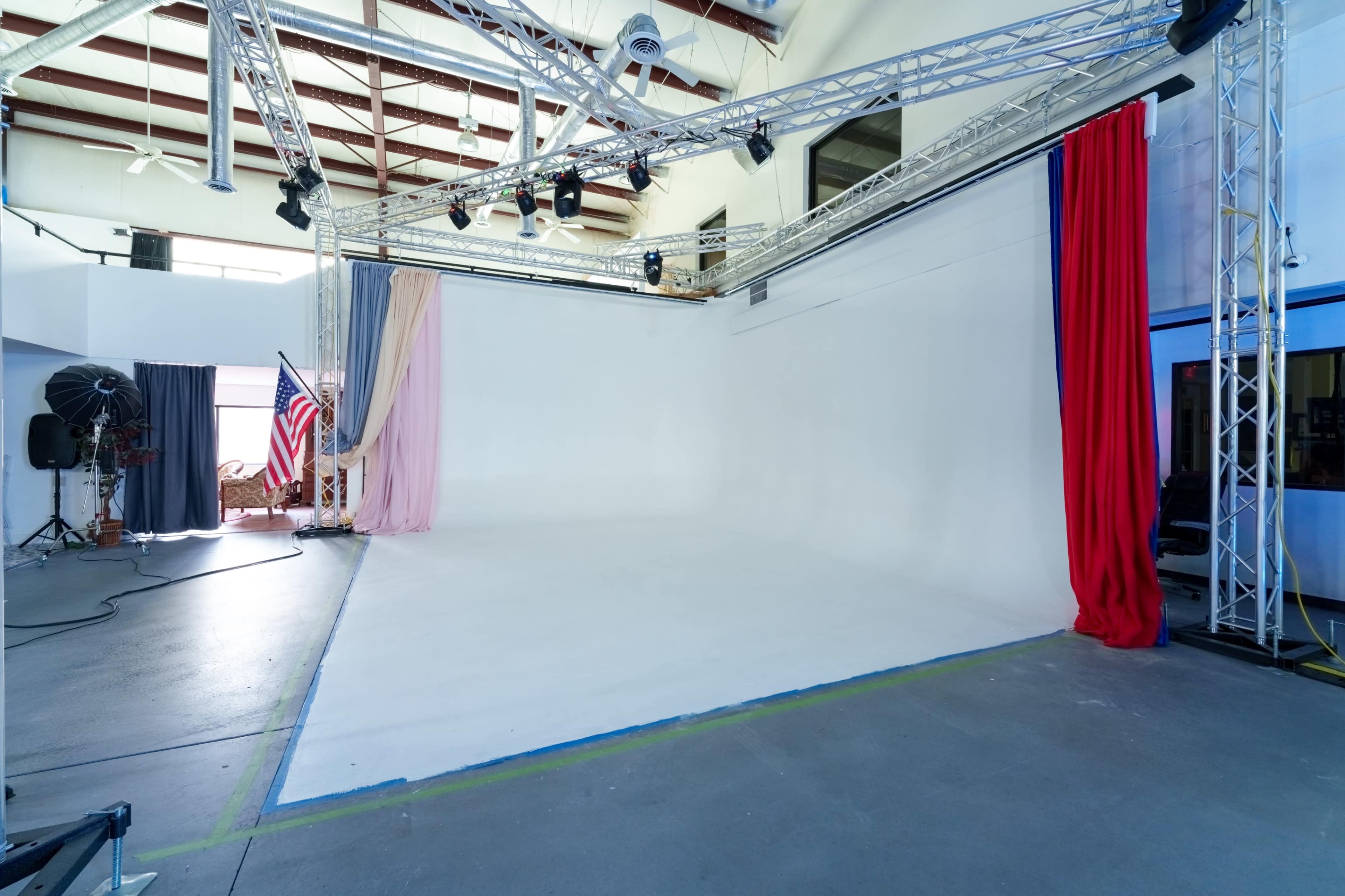 The image shows a photography studio with a large white backdrop, draped curtains in various colors, and lighting equipment scattered around.