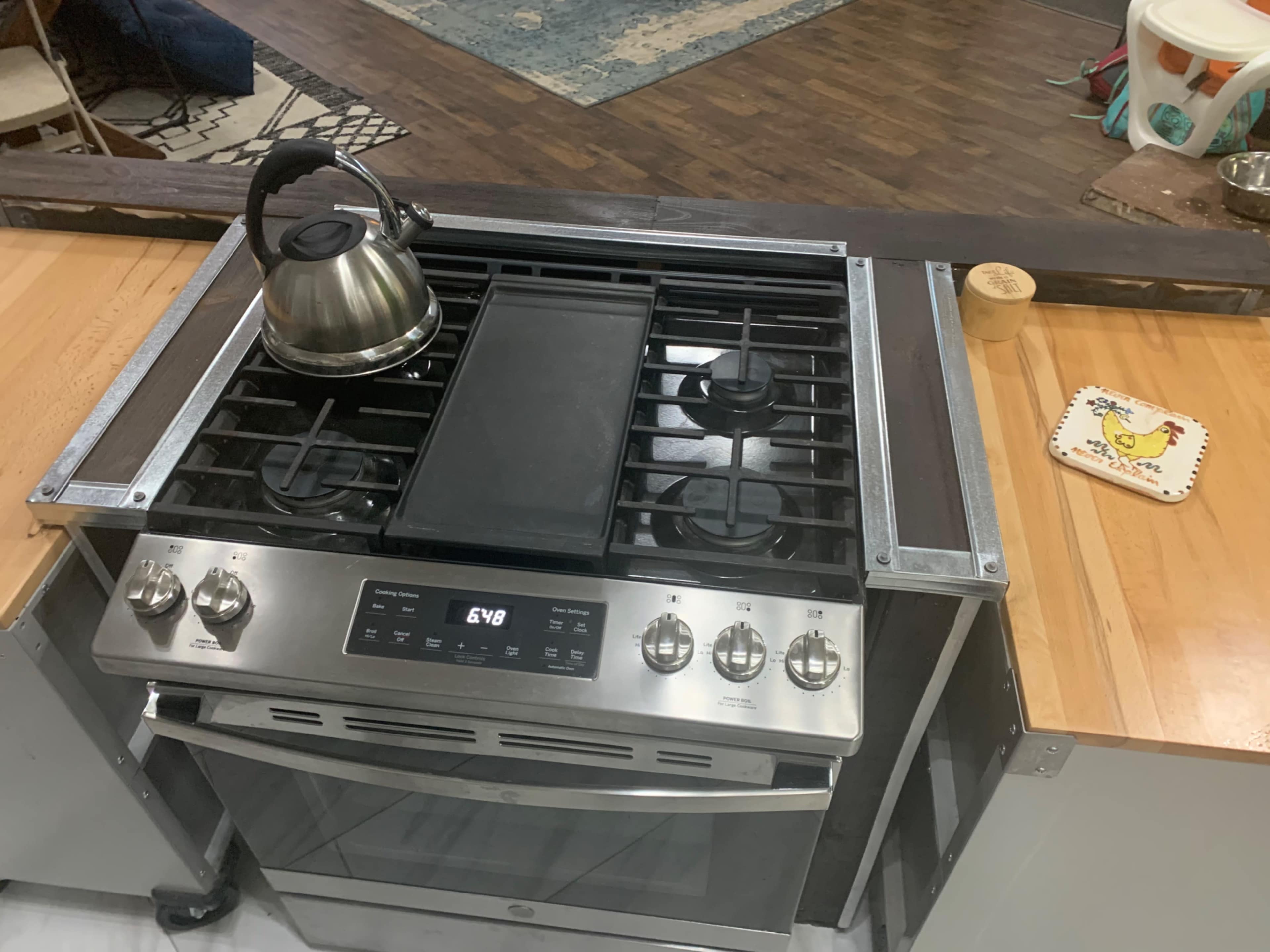 A stainless steel gas stove with a griddle and a kettle on top is positioned on a wooden countertop.