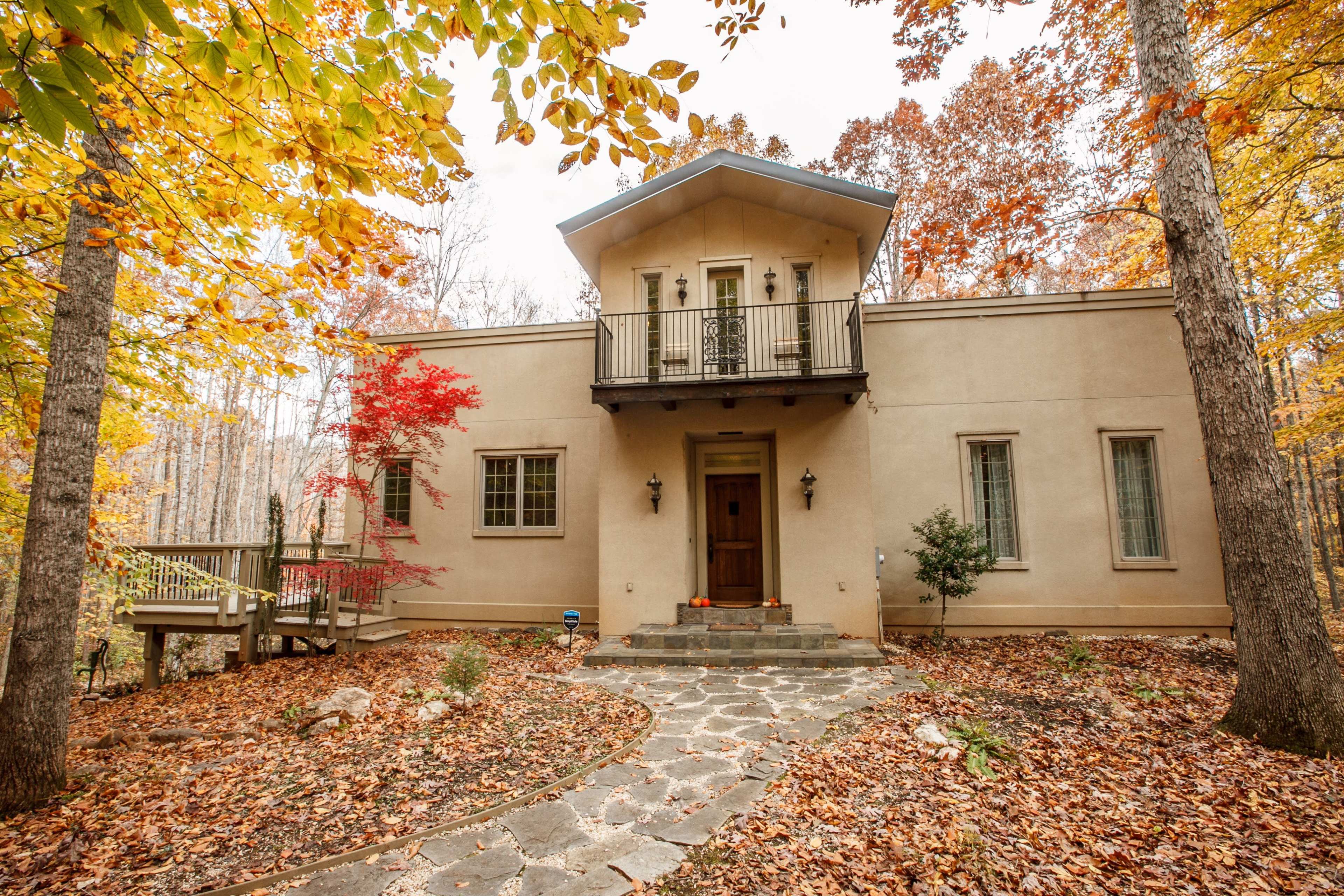 A modern house with a flat roof and a front porch is surrounded by trees with autumn foliage and a stone pathway leading to the entrance.