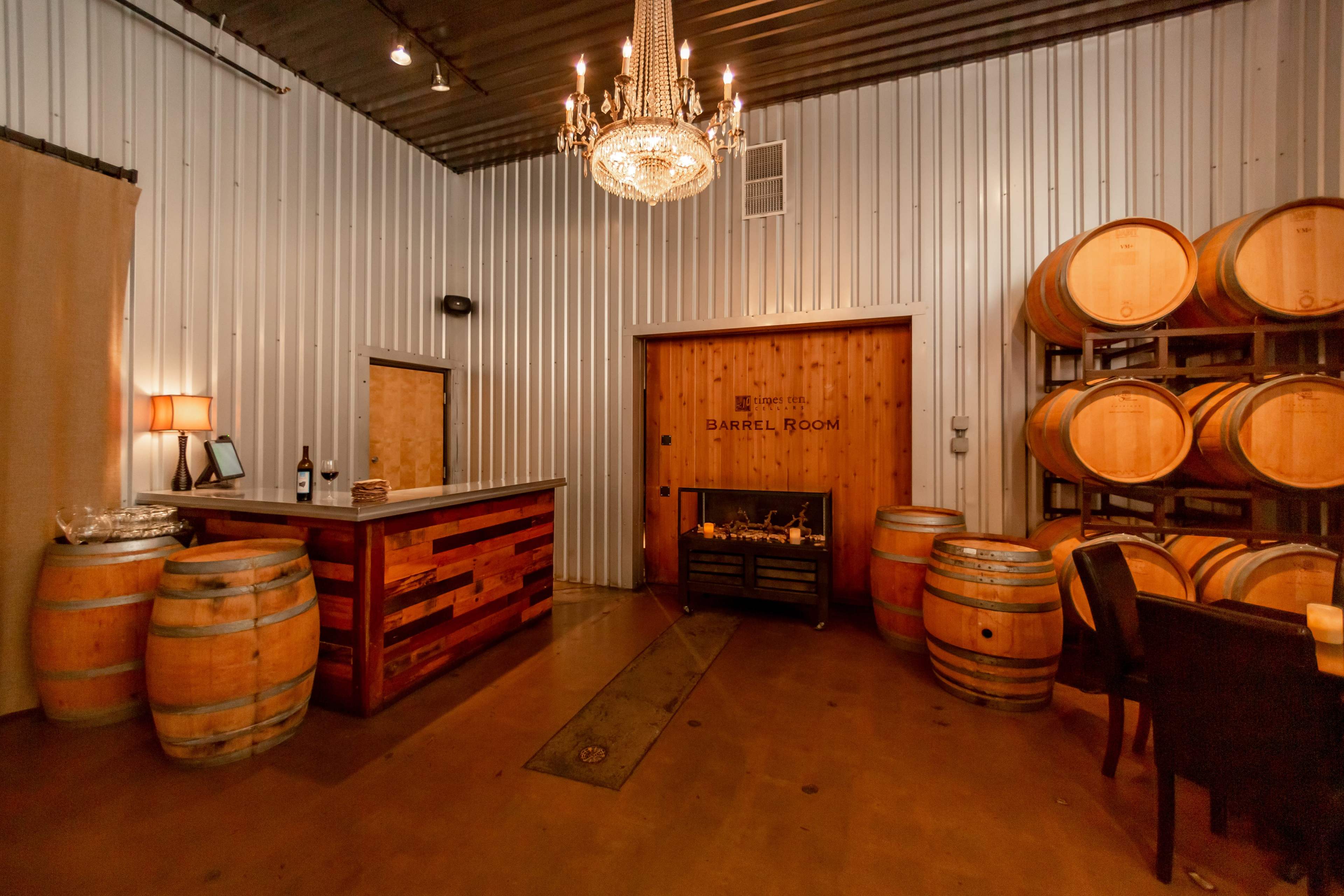 A winery tasting room featuring a wooden bar, wine barrels, and a chandelier against a backdrop of metallic walls.